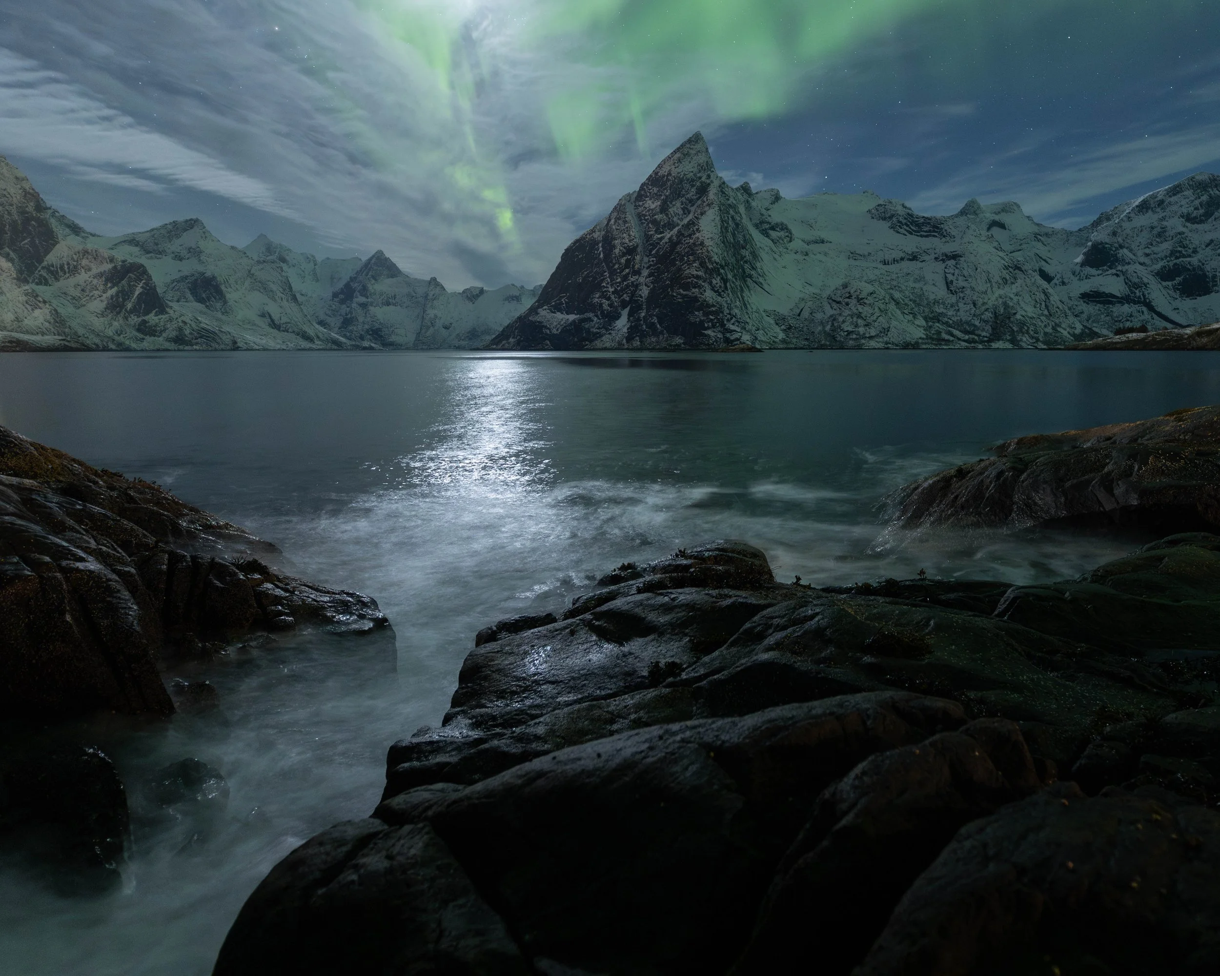 Olstinden cubierto de nieve, fiordo, rocas oscuras y aurora boreal iluminando el cielo sobre el paisaje ártico.