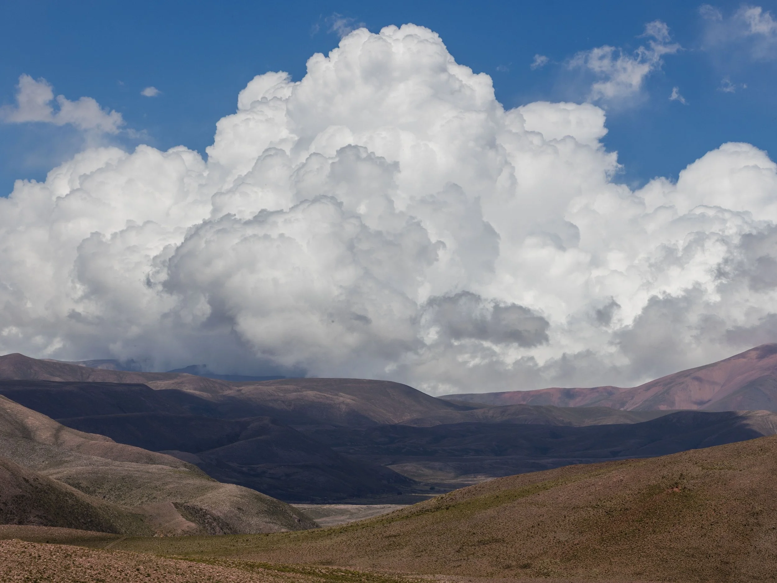 Fotografía de paisaje con enormes nubes blancas sobre montañas áridas, mostrando el contraste entre cielo azul, sombras profundas y relieve desértico en el norte de Argentina.