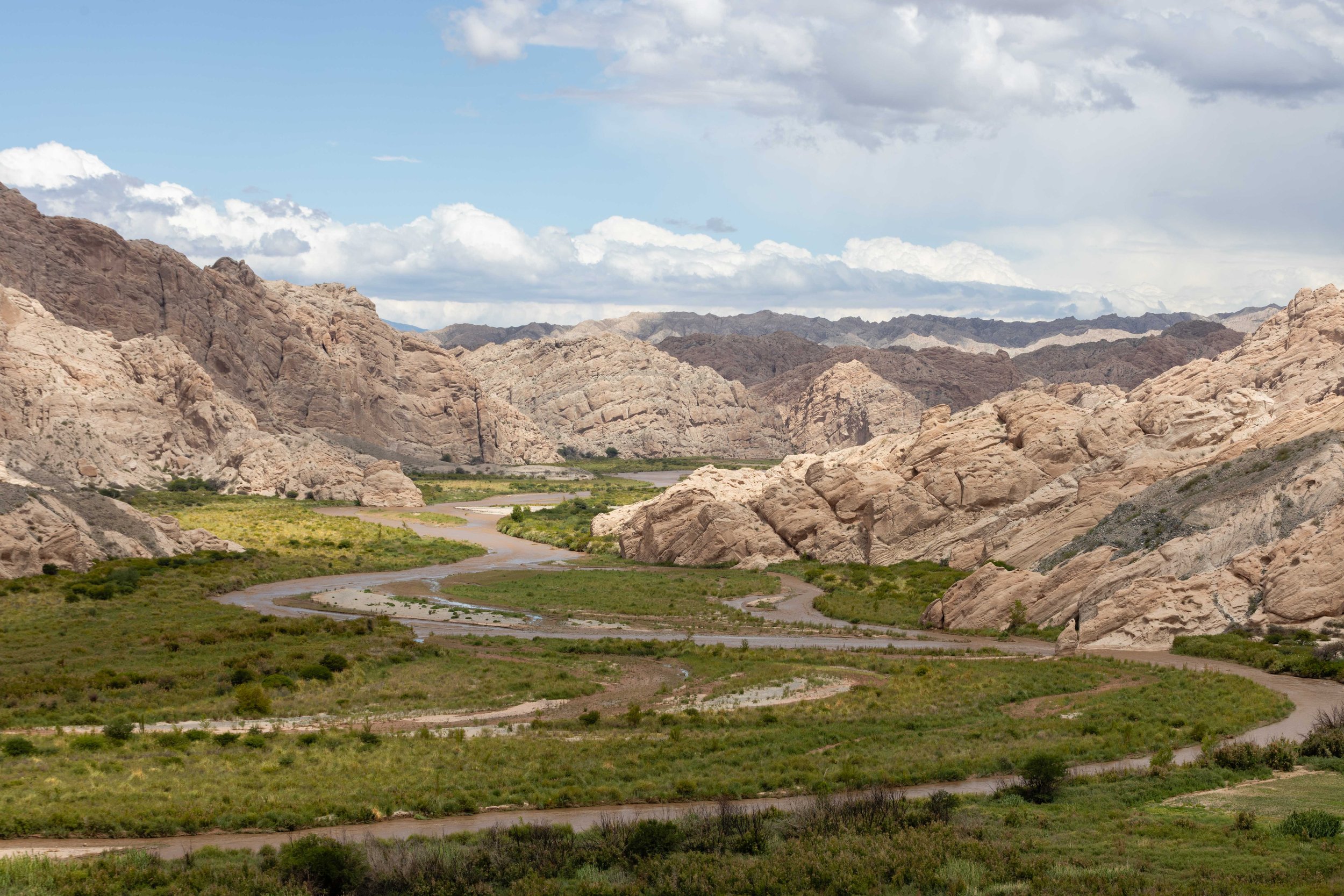 Fotografía de paisaje árido en el norte de Argentina, con un río atravesando un valle rodeado de formaciones rocosas, vegetación baja y cielo nublado.