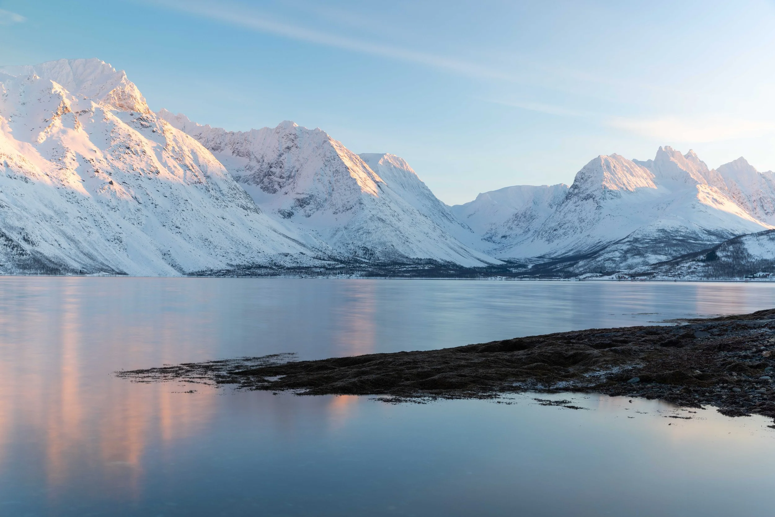 Fotografía de un fiordo noruego en invierno, con montañas cubiertas de nieve, reflejos delicados en el agua y luz pastel sobre el paisaje ártico. Alpes de Lyngen.