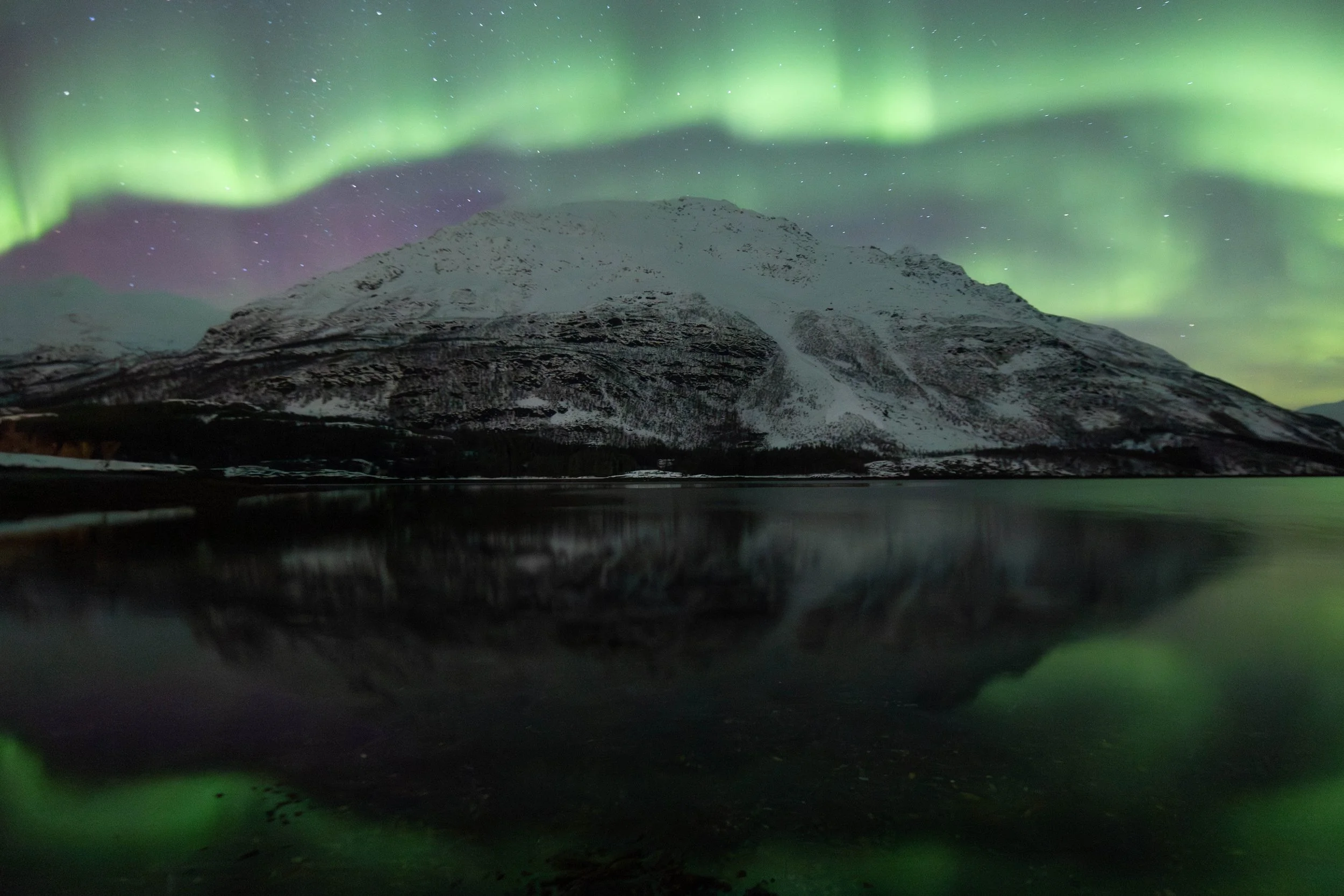 Fotografía de paisaje ártico con aurora boreal, montañas nevadas y reflejo sobre el agua, capturada durante una noche despejada en el norte de Noruega.