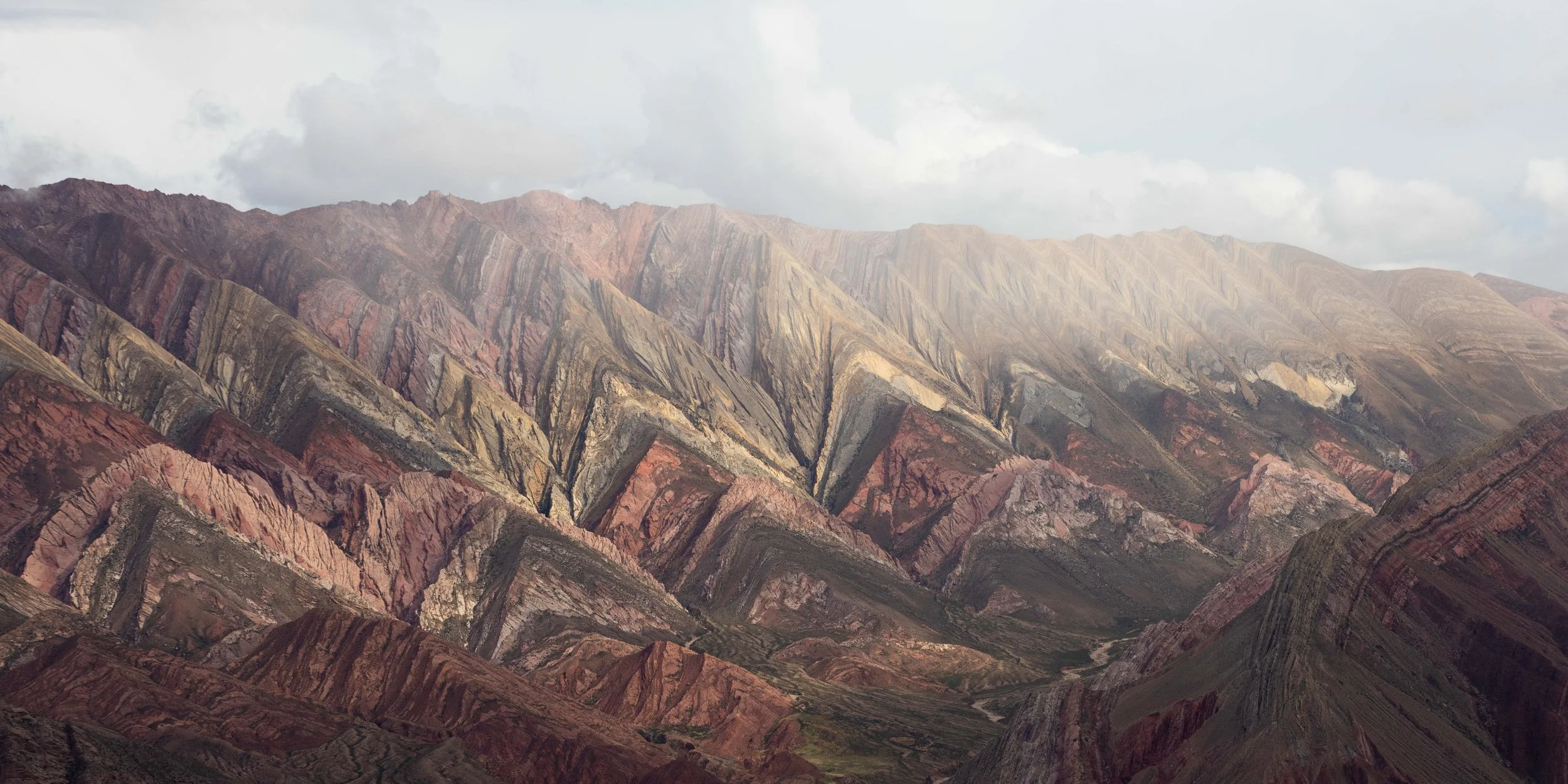 Fotografía panorámica del Hornocal  en el norte de Argentina, con capas geológicas marcadas, tonos rojizos y nubes suaves sobre el paisaje.