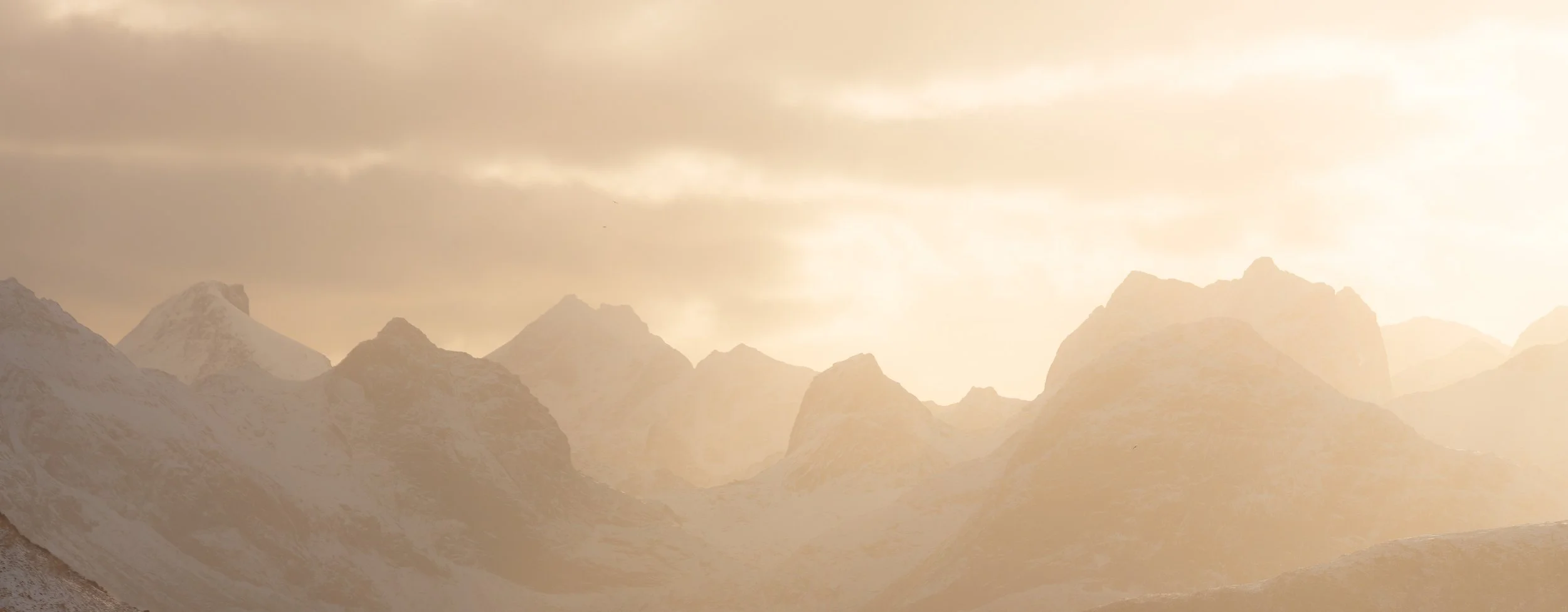 Imagen panoramica de montañas en Lofoten norte de Noruega con luz cálida atravesando las nubes, creando una atmósfera suave, nevada y contemplativa.