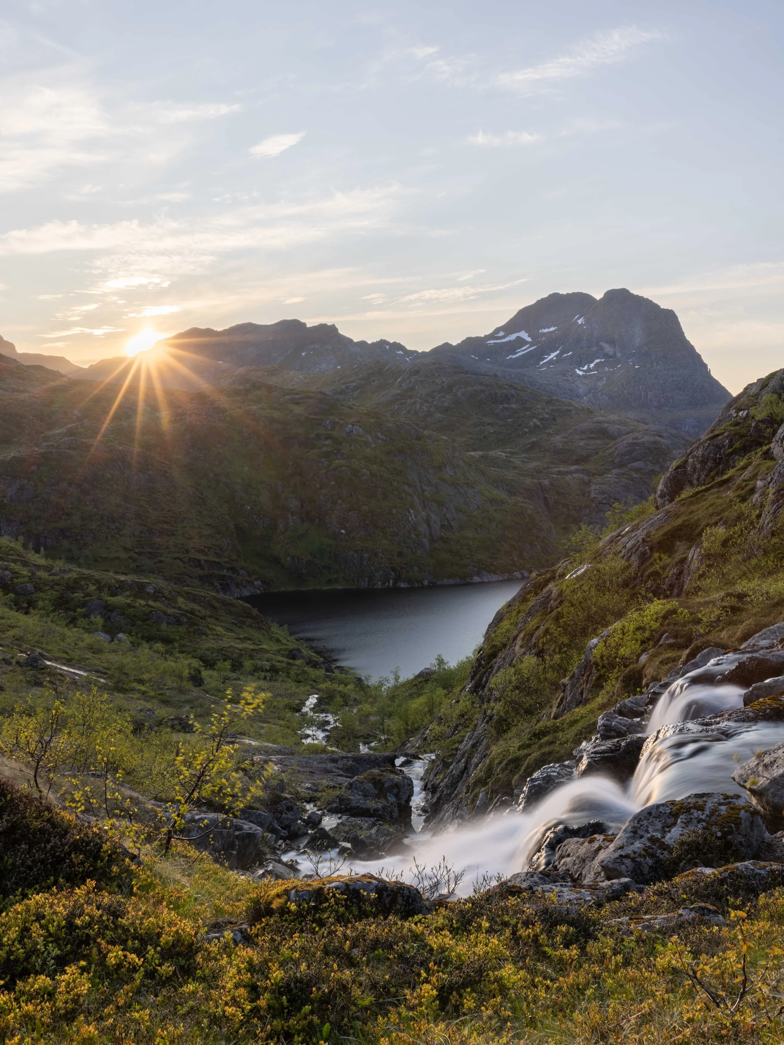 Paisaje de Lofoten con cascada, montañas, lago y luz cálida del sol de medianoche, capturando la belleza salvaje del verano en el norte de Noruega.