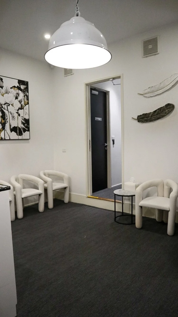 A waiting room with white chairs, a small round table with tissue, a large white ceiling lamp, black and white wall art, and a door labeled 'Toilets'.