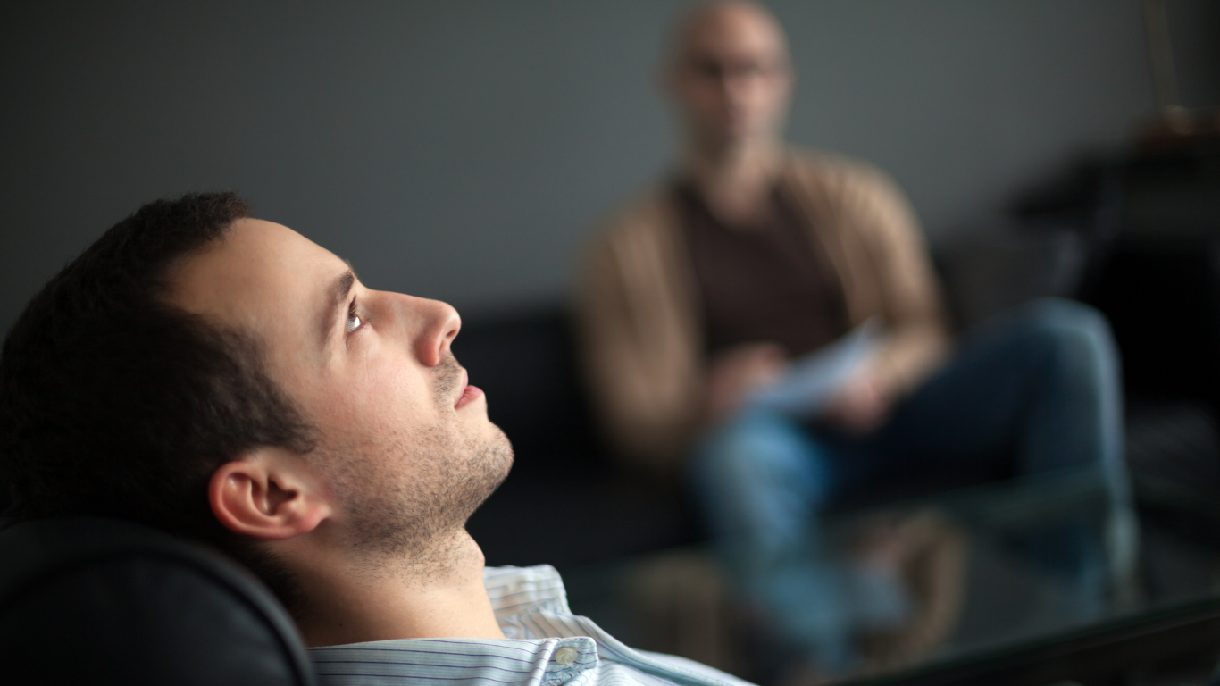 A man looking up and to the side while sitting in a therapy or counseling session with a blurred therapist in the background.