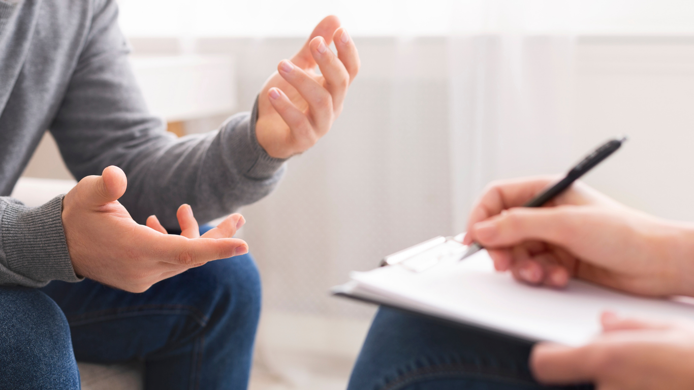 Two people having a conversation during a counseling or therapy session, with one person taking notes on a clipboard.