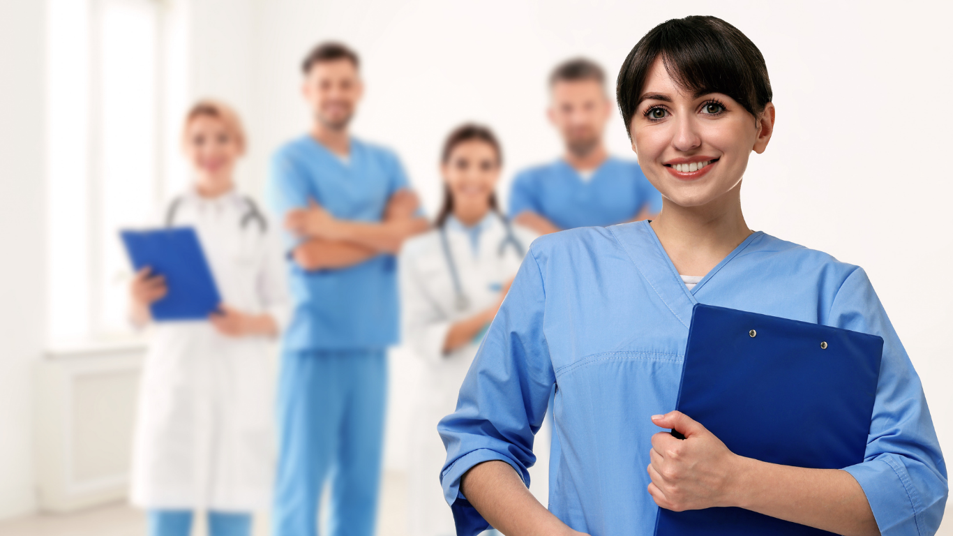 Healthcare professionals, including nurses and doctors, standing together in a white room, with the woman in the foreground smiling and holding a blue clipboard.