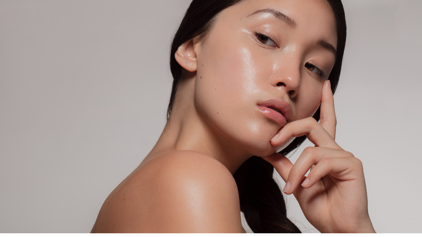 Close-up of a woman with smooth, glowing skin touching her face, with a neutral background.