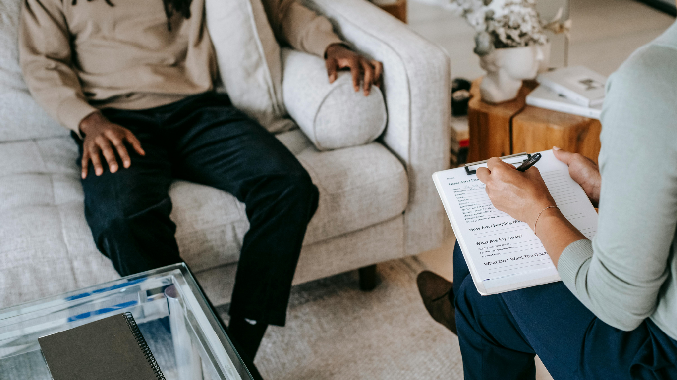 A person sitting on a beige sofa, facing a therapist who is taking notes on a clipboard in a therapy or counseling session.