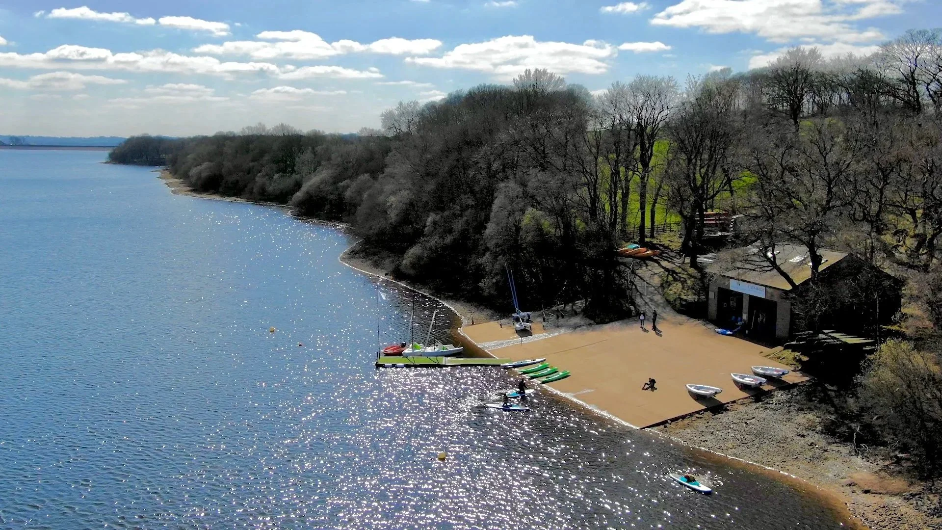 A lakeside with small sailboats, kayaks, and paddleboards on a sandy beach, surrounded by trees with some leaves, under a partly cloudy sky.