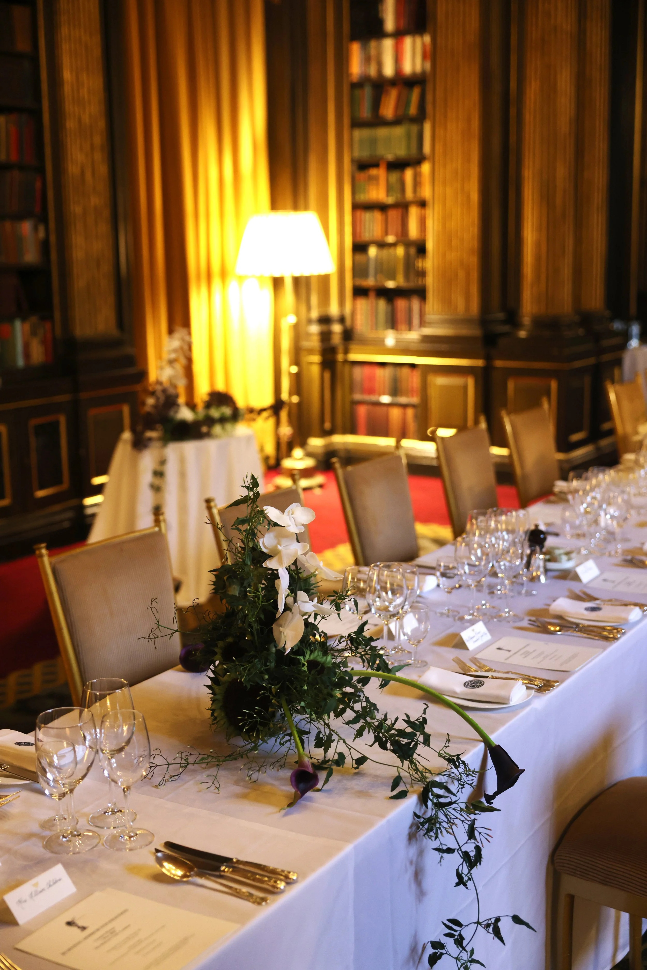 Elegant dining table set for a formal event with white tablecloths, glassware, silverware, and floral centerpieces, in a room with wooden bookshelves and warm lighting.