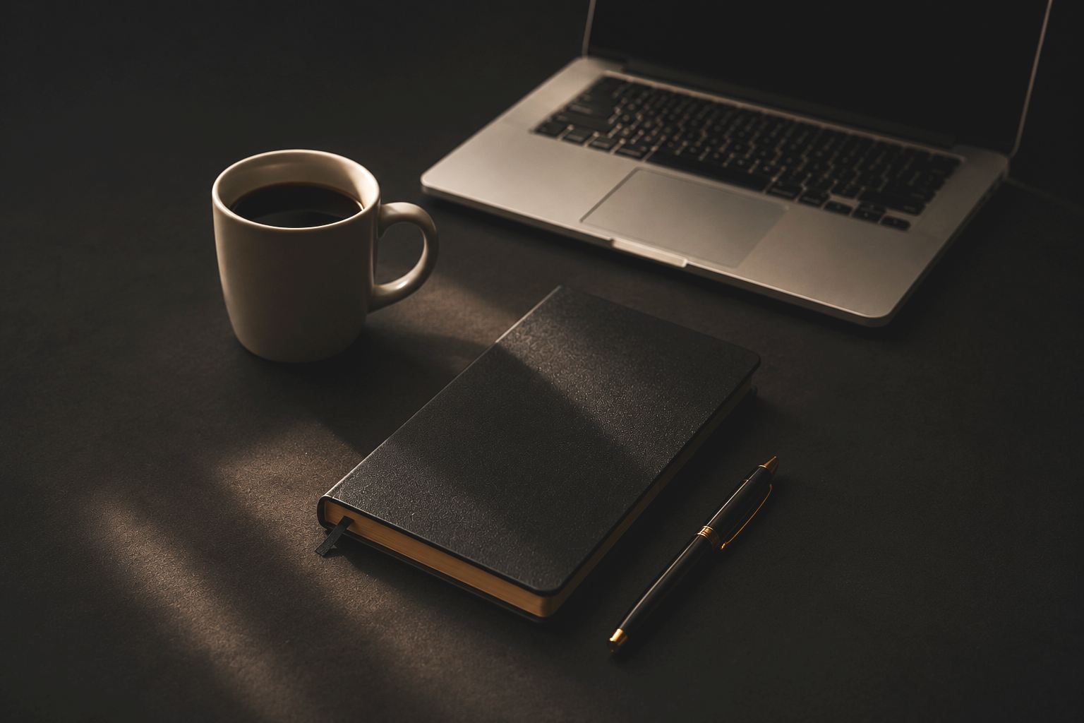 A desk with a white coffee mug filled with black coffee, a closed black notebook, a silver laptop with an open keyboard, and a black and gold pen on a dark surface.