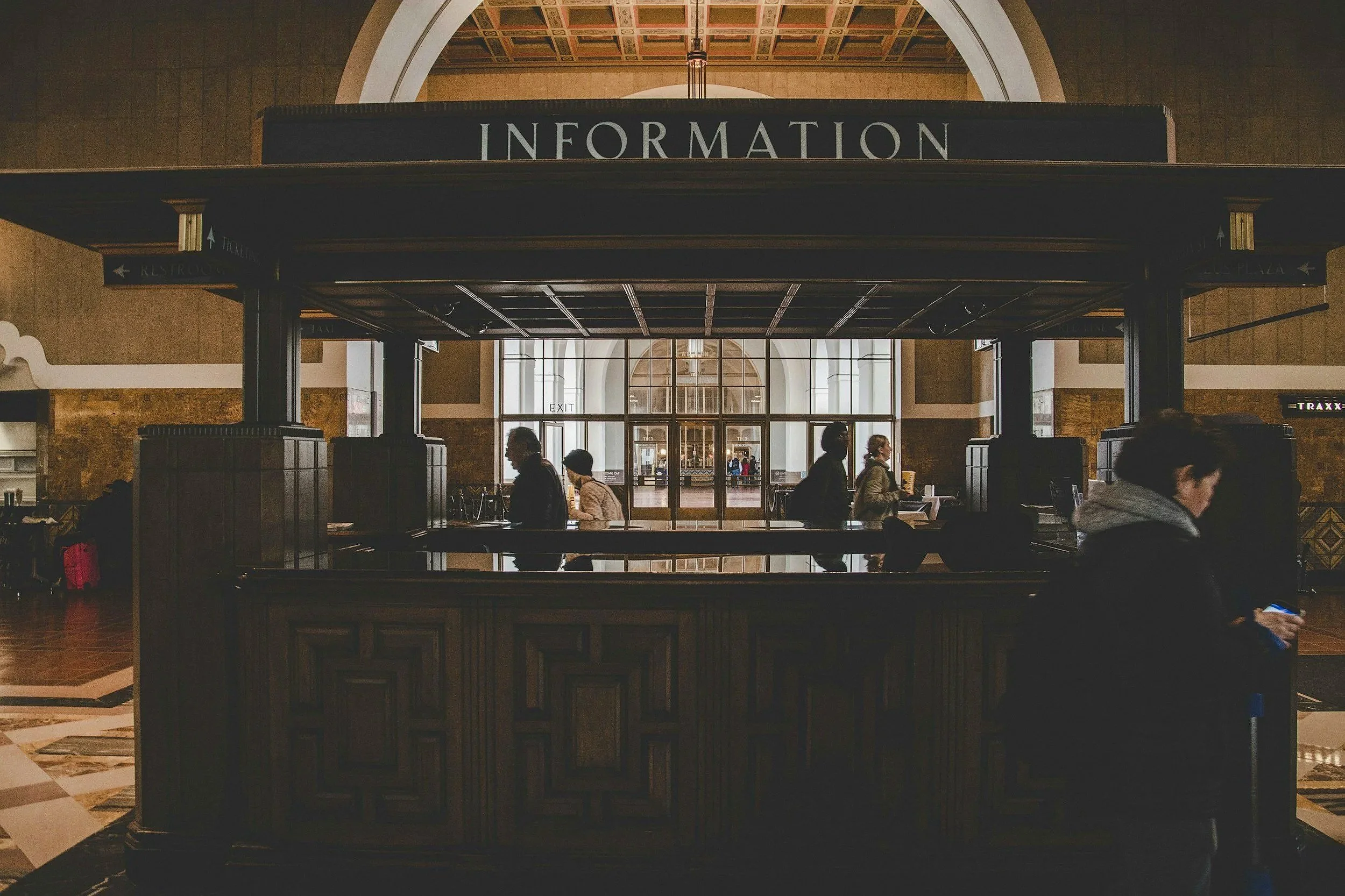 Large black information booth at an airport terminal with people walking around and through a glass door behind it; above the booth, a sign reads 'INFORMATION'; the ceiling features ornate detailing.