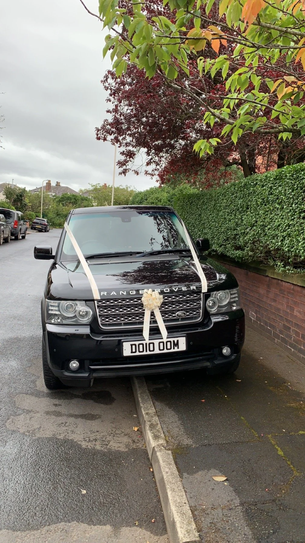 Black Range Rover parked on the side of a street decorated for a celebration, with a large cream-colored ribbon and bow on the hood, and a custom license plate that reads 'DOIO OOM'.