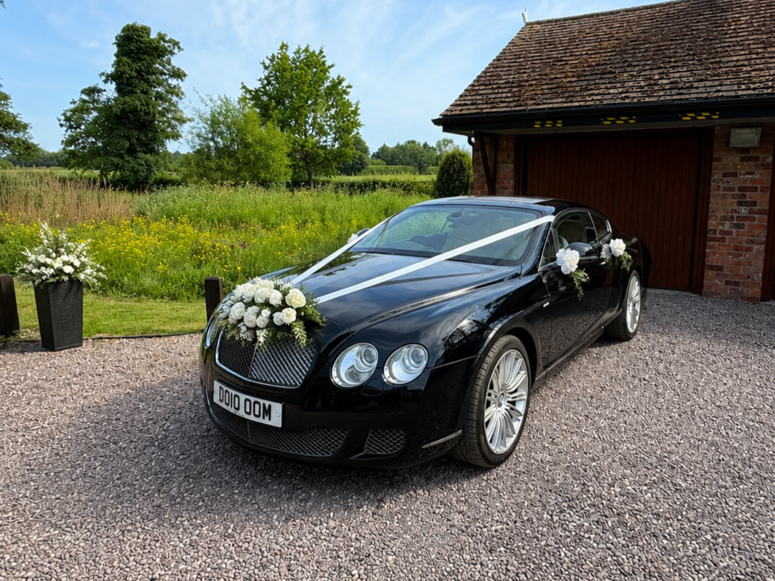 Black luxury Bentley decorated with white flowers and ribbons, parked on gravel driveway outside a brick house with a red wooden door, greenery and trees in the background.
