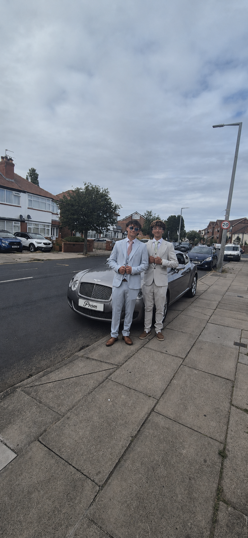 Two young men in formal suits standing in front of a luxury car with a sign that reads 'Prom', on a suburban street with parked cars and houses in the background.