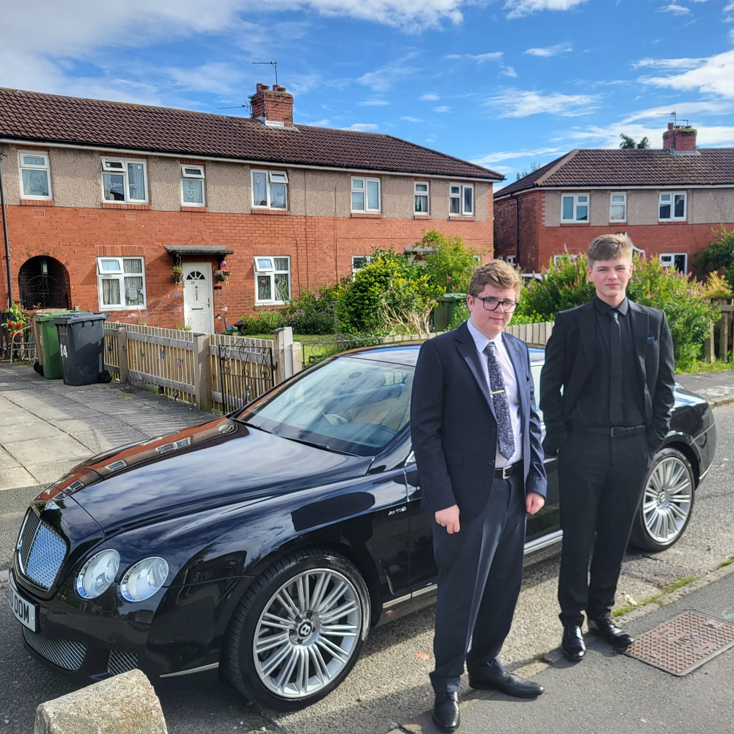 Two young men in suits standing next to a black luxury car parked on the street in front of a row of houses. The houses have brick and stucco exteriors with white-framed windows and red tiled roofs. The scene is outdoors during daytime with a blue sky and some clouds.