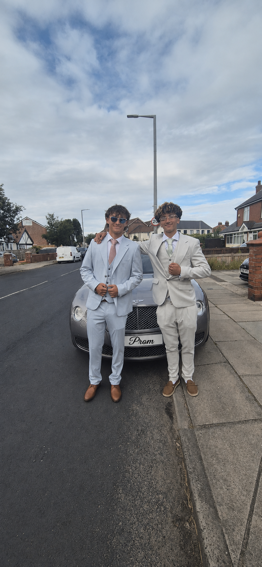 Two young men dressed in suits stand in front of a sports car on a suburban street. The man on the left wears a light gray suit with a tie and sunglasses. The man on the right wears a cream suit and is making a fist while smiling. The car has a personalized license plate that says 'Prom.' The sky is partly cloudy, and there are houses and cars in the background.