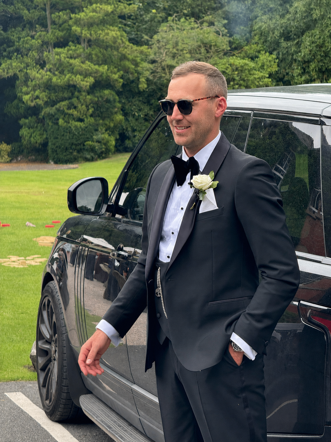 A man in a tuxedo with sunglasses and a white rose boutonniere standing next to a black SUV outdoors with trees and grass in the background.
