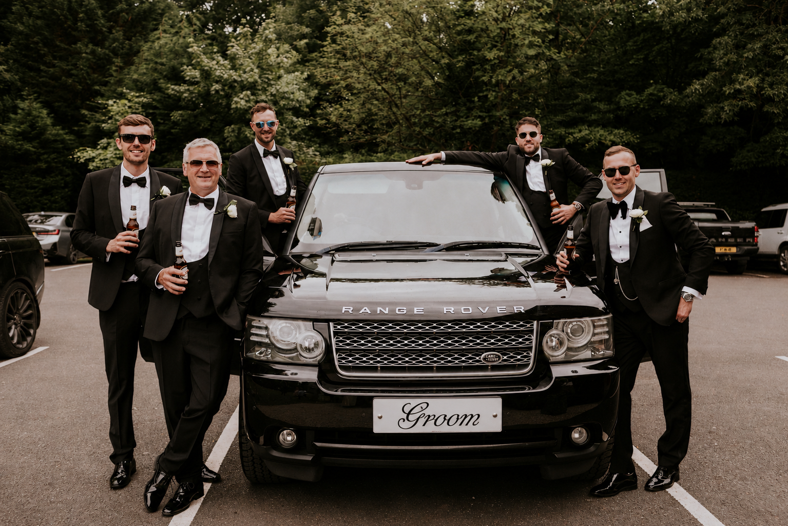Six men dressed in tuxedos, some wearing sunglasses, posing with a black Range Rover in a parking lot, with a green forested background. They are smiling, holding beer bottles, and one man is leaning on the car's roof. A custom license plate reads 'Groom'.