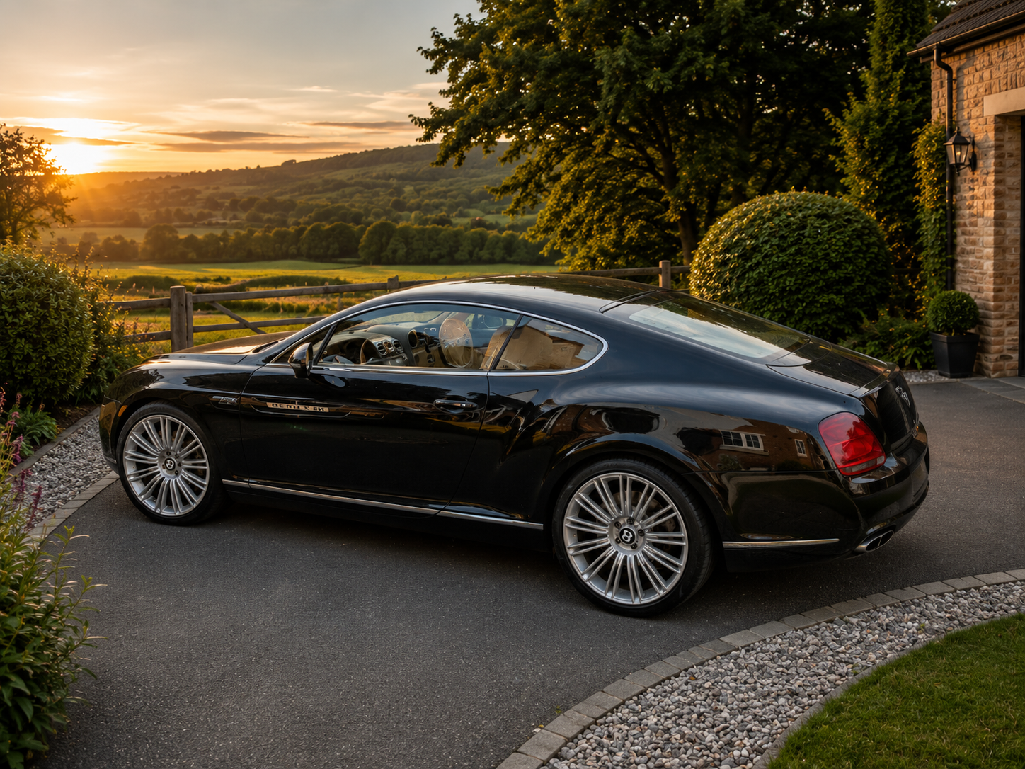 A black luxury car parked in a driveway during sunset, with lush green trees and rolling hills in the background.
