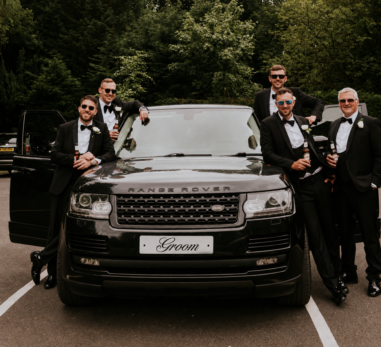 Six men in tuxedos and sunglasses are standing around and sitting in front of an all-black Range Rover with a 'Groom' sign on the front license plate. They are holding beer bottles and smiling, in a parking lot with trees in the background.