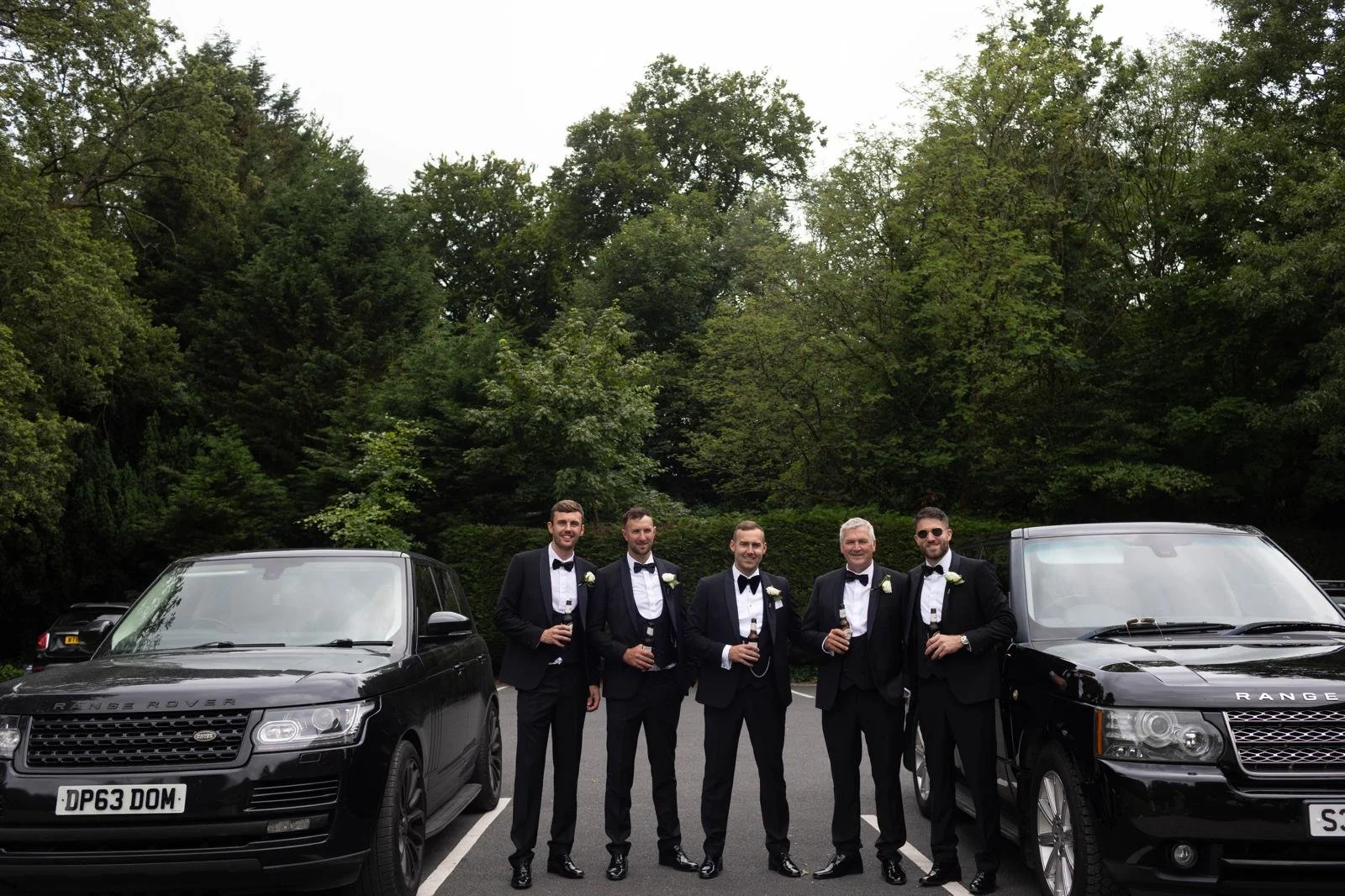 Group of five men in tuxedos standing between two black Range Rover vehicles in a parking lot surrounded by green trees.