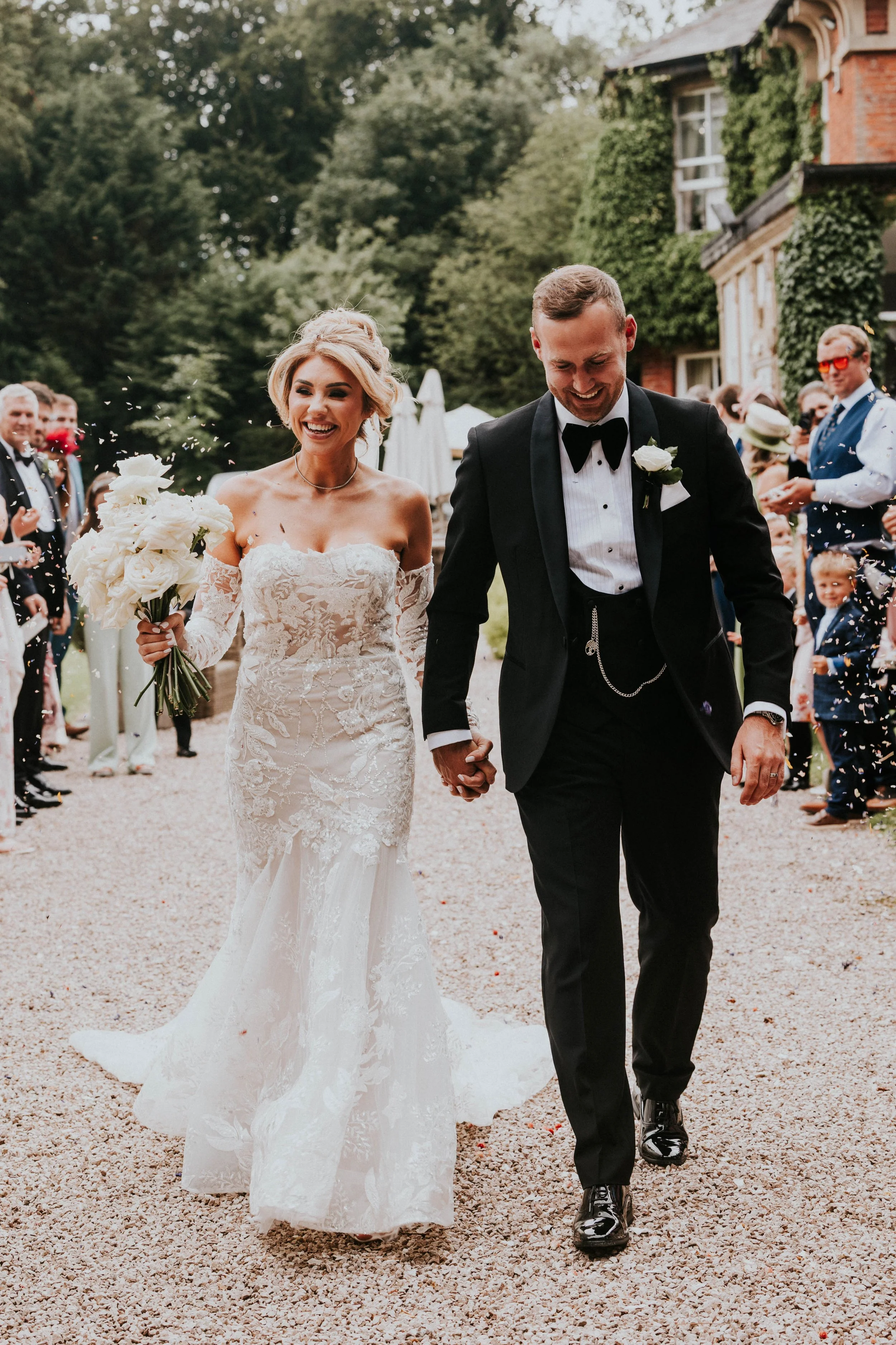 A bride and groom walking hand in hand outdoors after their wedding ceremony, surrounded by guests and greenery.