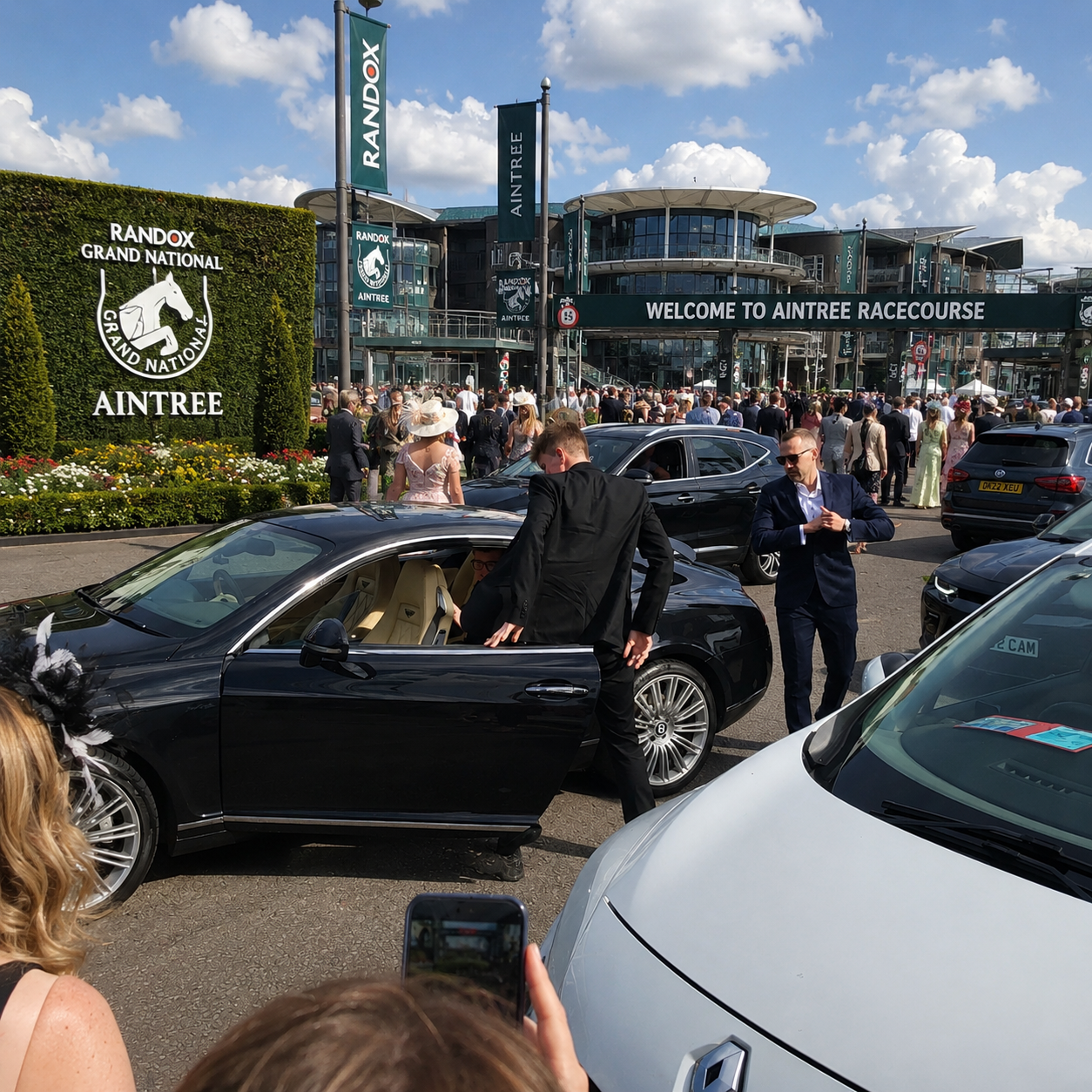 Crowded scene outside Aintree Racecourse with people dressed in formal attire, luxury cars, and a large sign welcoming visitors to Aintree Racecourse.