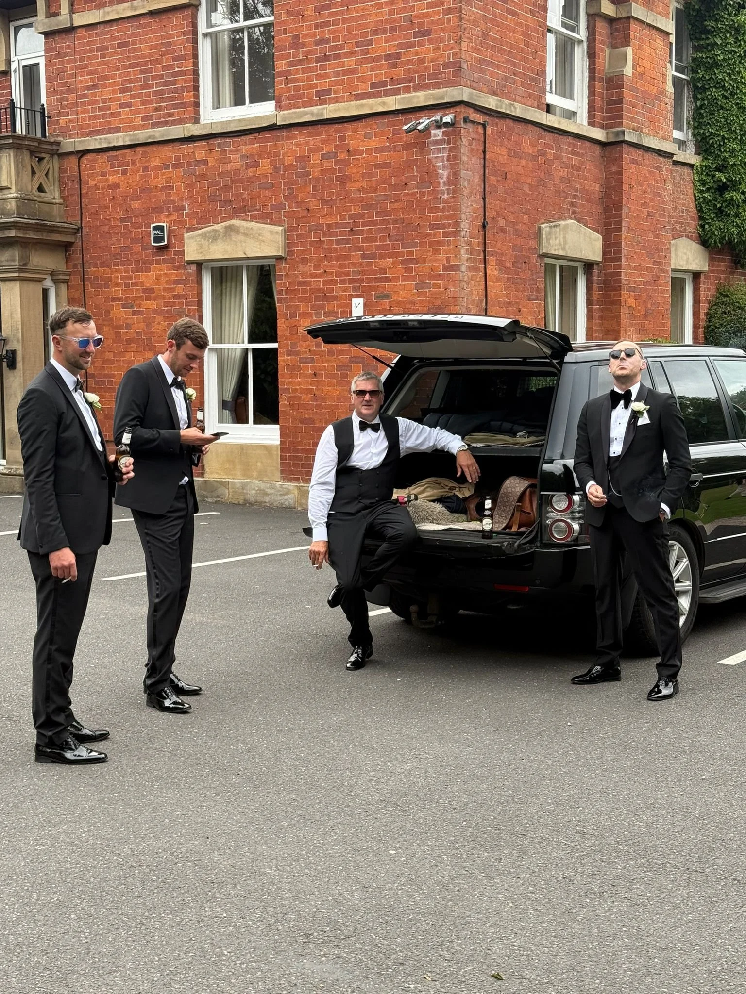 Four men dressed in tuxedos standing outside near a black SUV with its trunk open, in front of a red brick building.