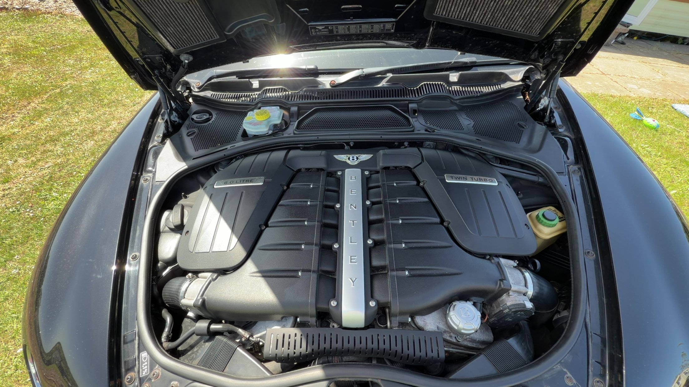 View of a car engine with the hood open, featuring a Bentley engine labeled '6.0 Litre' and 'Twin Turbo' on the cover, located outdoors on a grassy area.