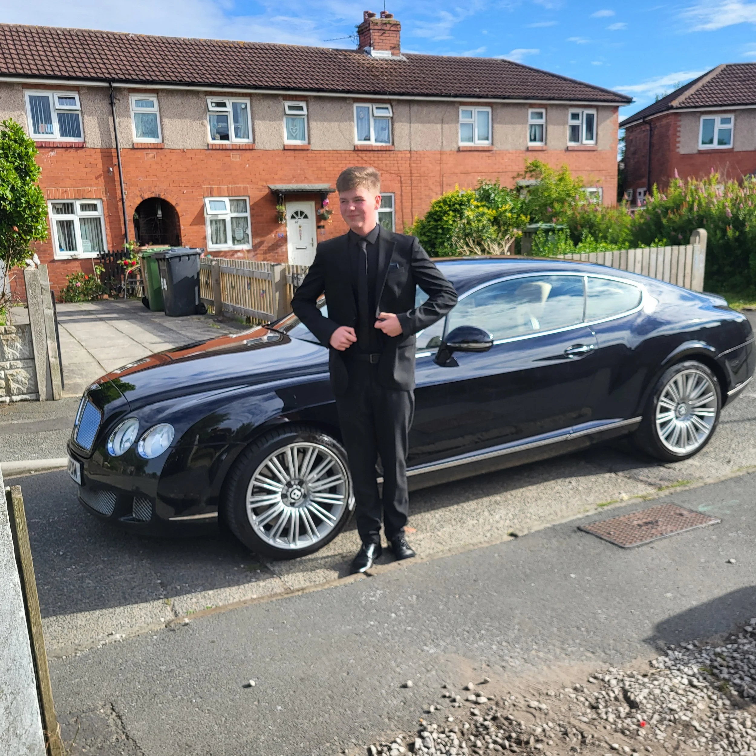 A young man dressed in a black suit standing outside in front of a black luxury car, with old brick houses and a garden in the background.