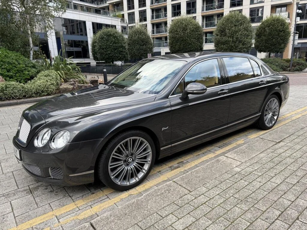 A black luxury Bentley parked on a city street, with modern apartment buildings and landscaped greenery in the background.