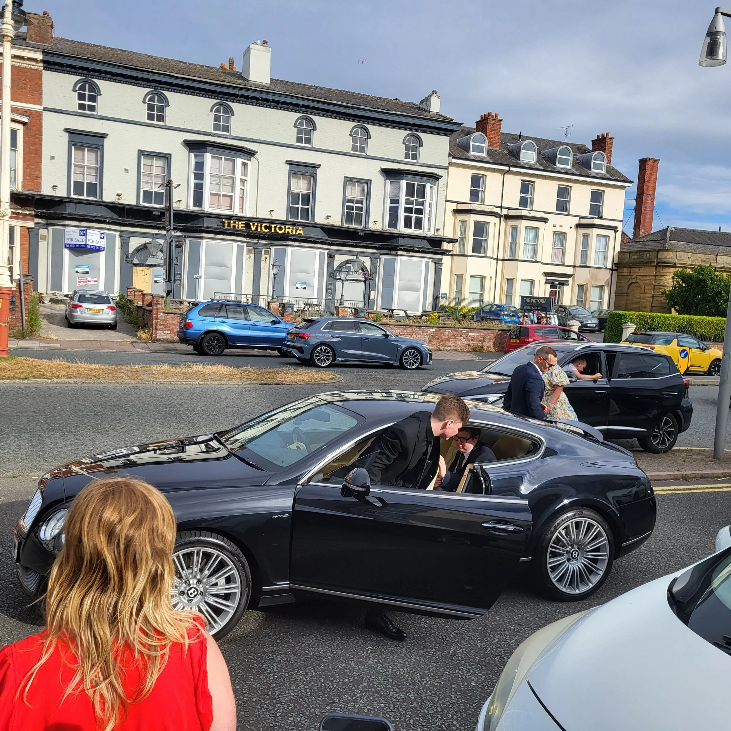 A parking lot in front of a building called 'The Victoria' with several cars and people, including a woman with red hair and a man in a black jacket looking into a car.
