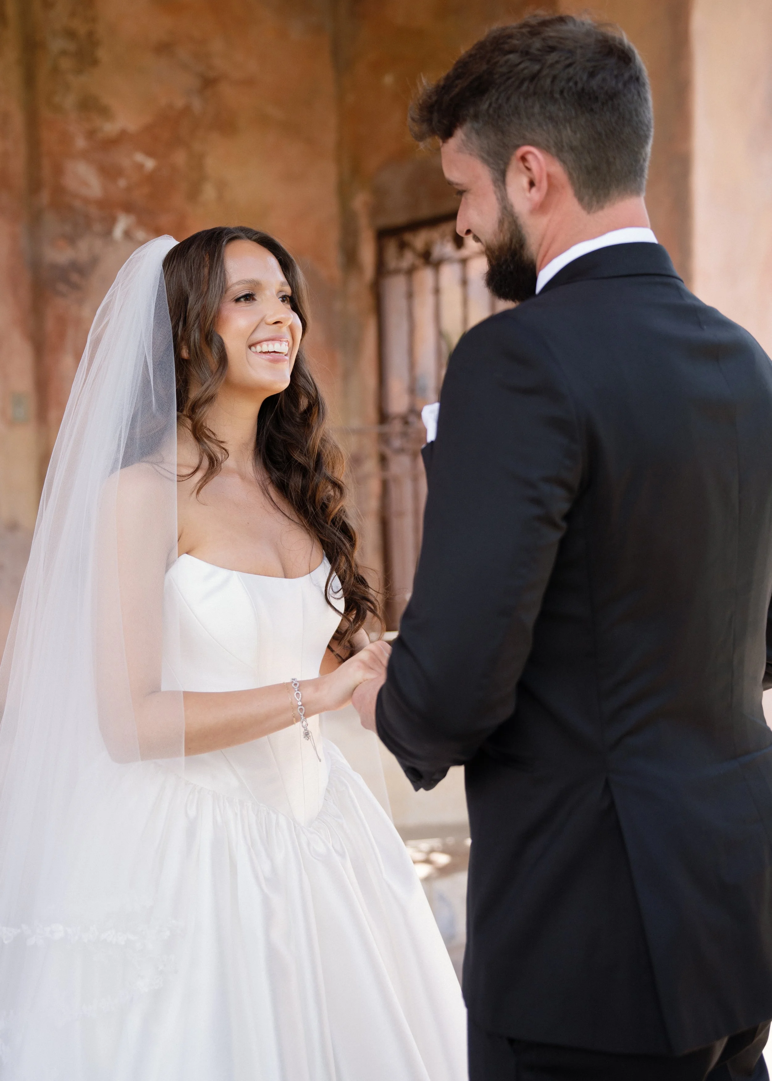 A bride and groom holding hands during their wedding ceremony. The bride is smiling and wearing a white wedding gown with a veil, while the groom is dressed in a black suit.