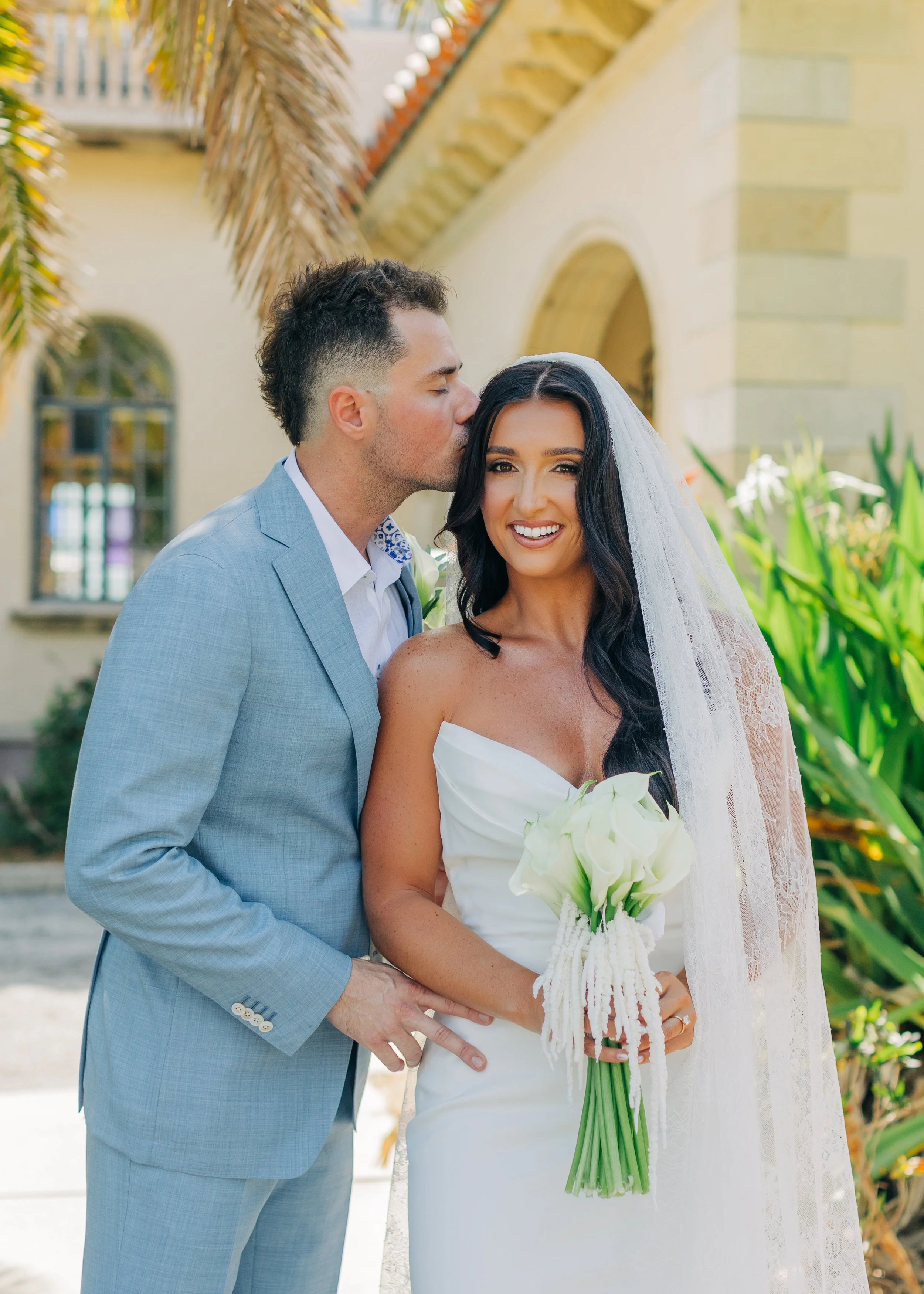 A newlywed couple with the groom kissing the bride on the temple, in an outdoor setting with greenery and a building in the background. The bride holds a bouquet of white calla lilies and wears a white wedding dress with a lace veil.