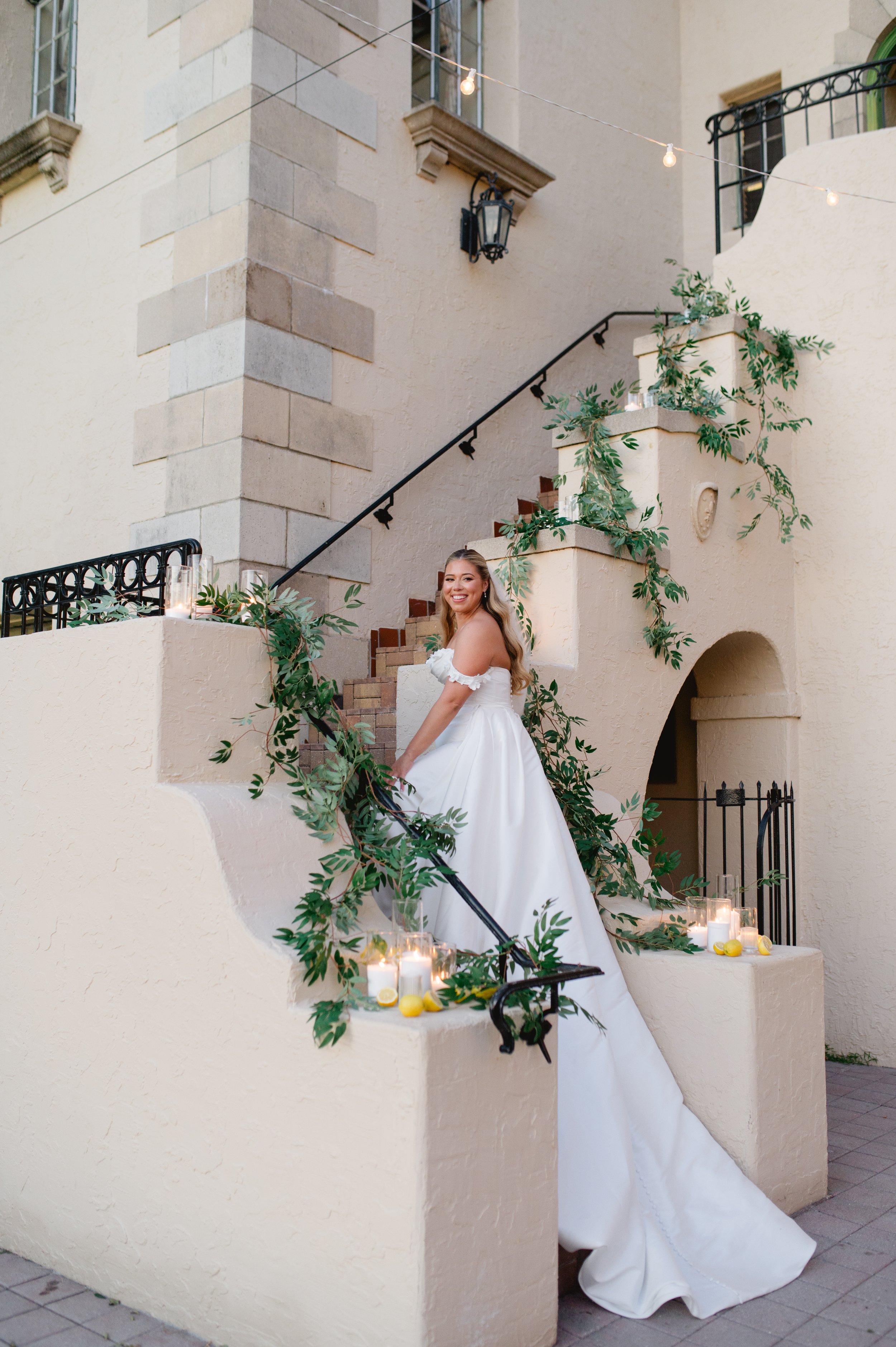 A woman in a white dress smiling while standing on a decorated outdoor staircase with candles, green foliage, and a yellow lemon, against a beige stucco building with windows and a wrought iron railing.