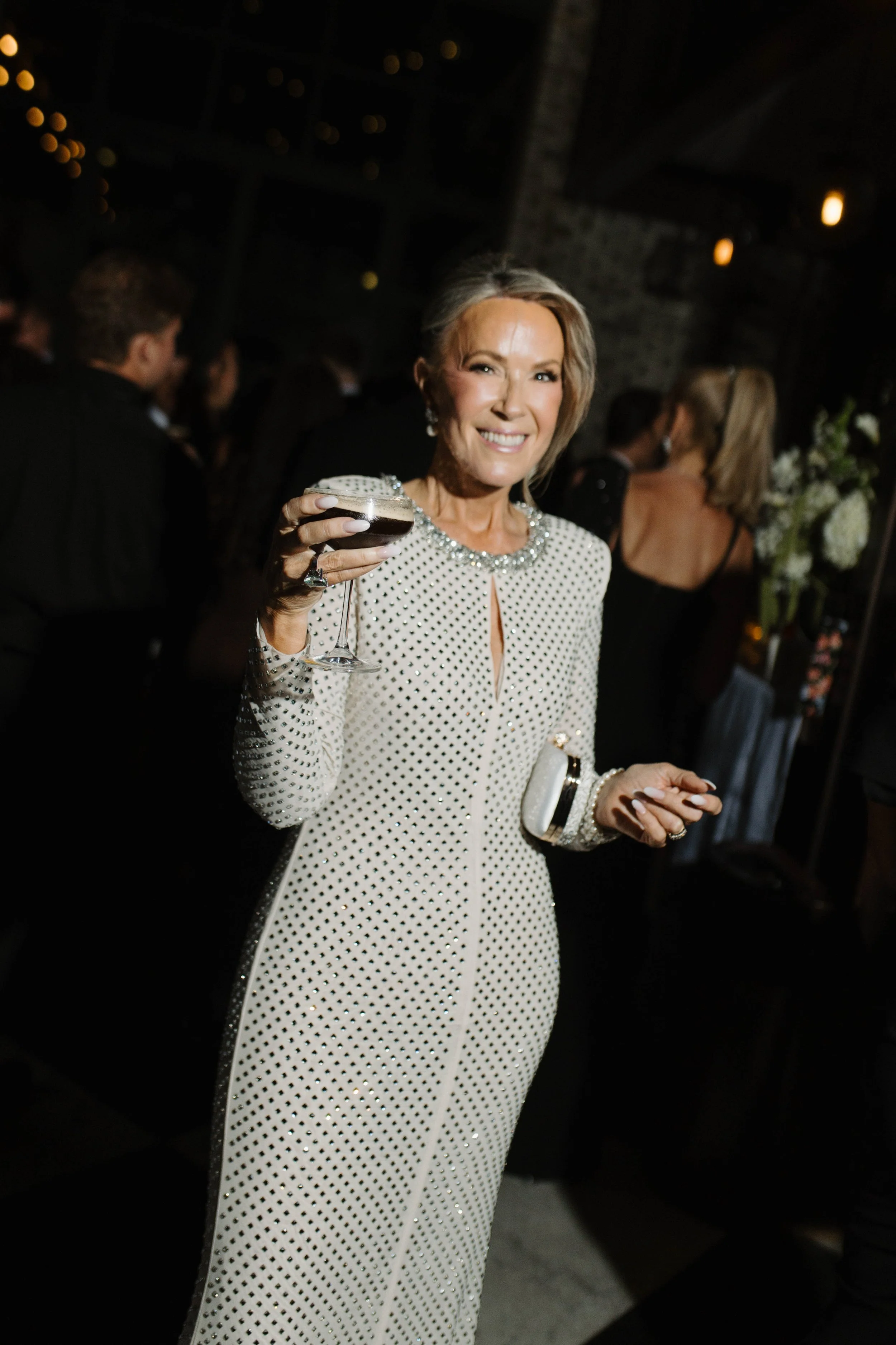 A smiling woman in a white, dotted dress holding a cocktail at a social gathering or party.