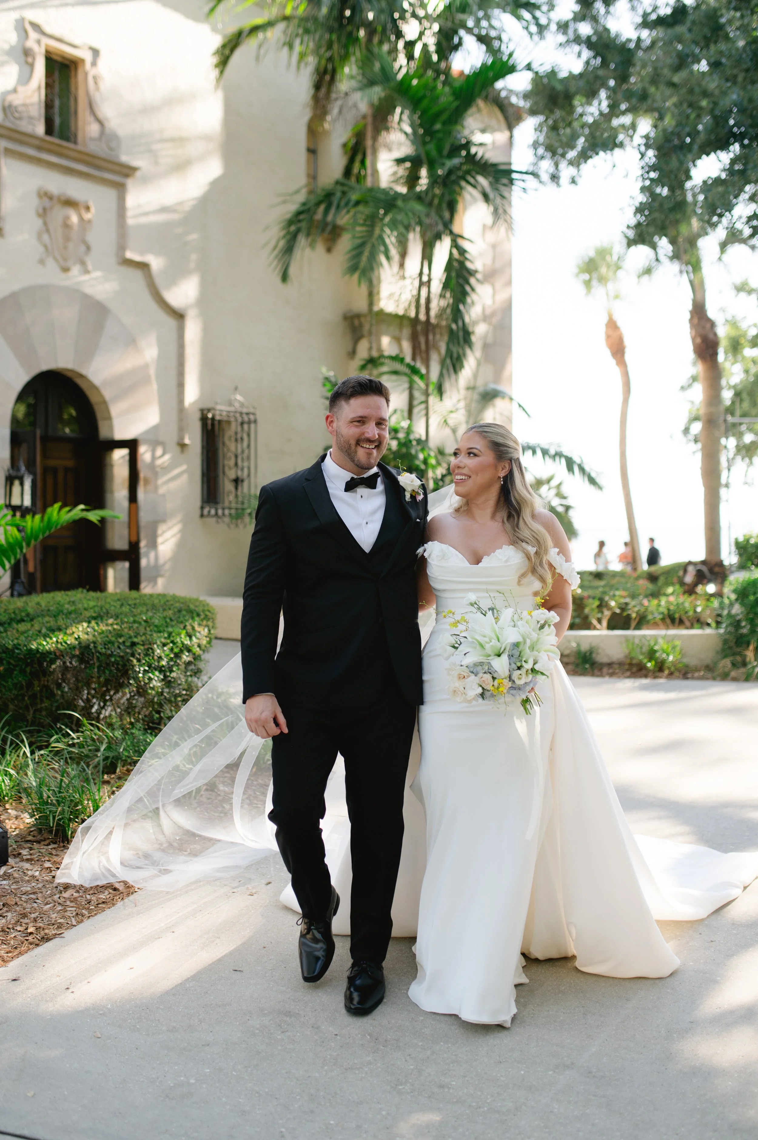 A happy bride and groom walking outdoors in a garden, with the bride holding a bouquet of white lilies and wearing a white wedding dress, and the groom in a black tuxedo with a bow tie, surrounded by lush green trees and a historic building in the ba