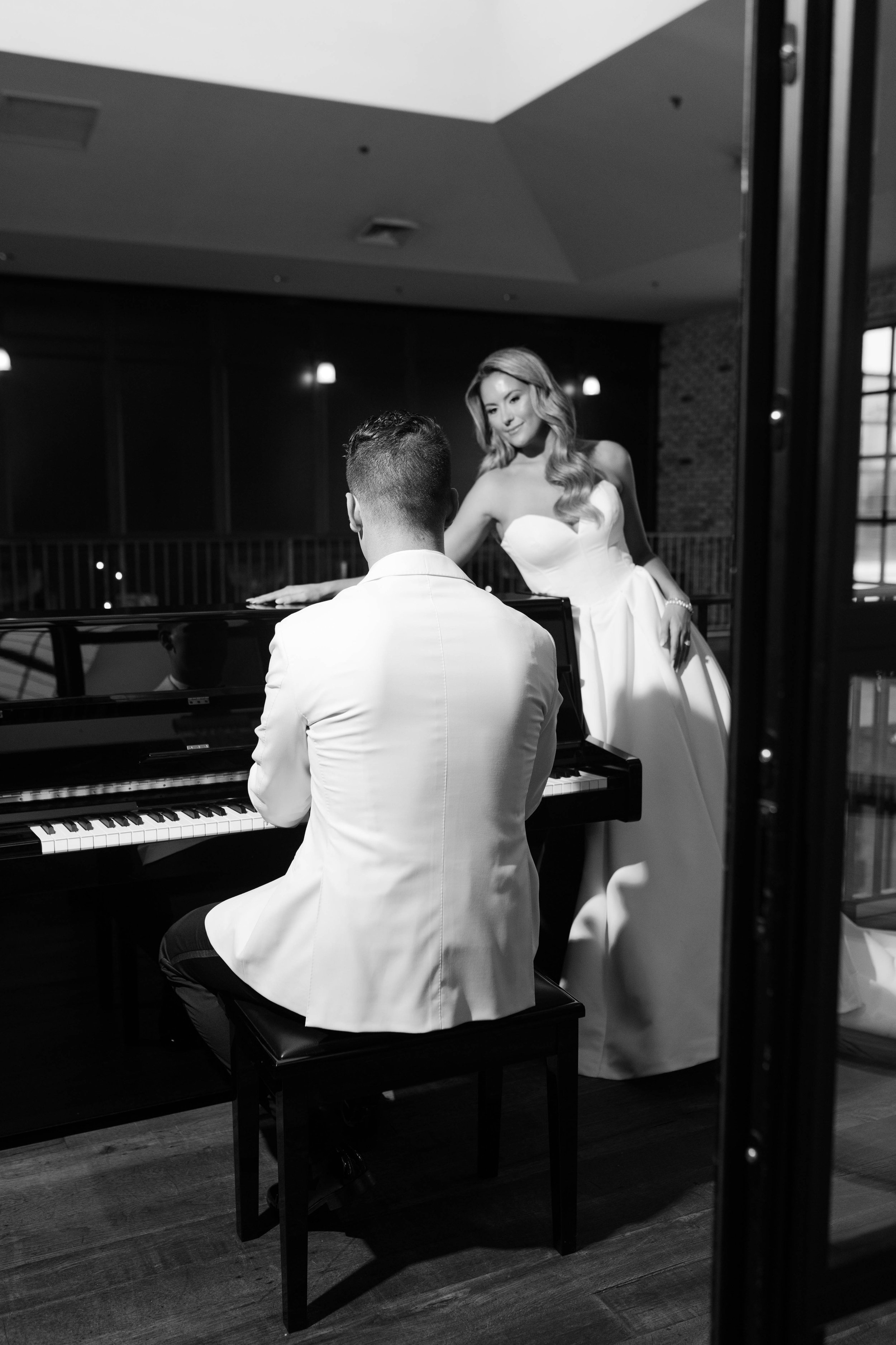 A woman in a strapless white gown stands beside a man playing the piano, both in a dimly lit room with dark walls.