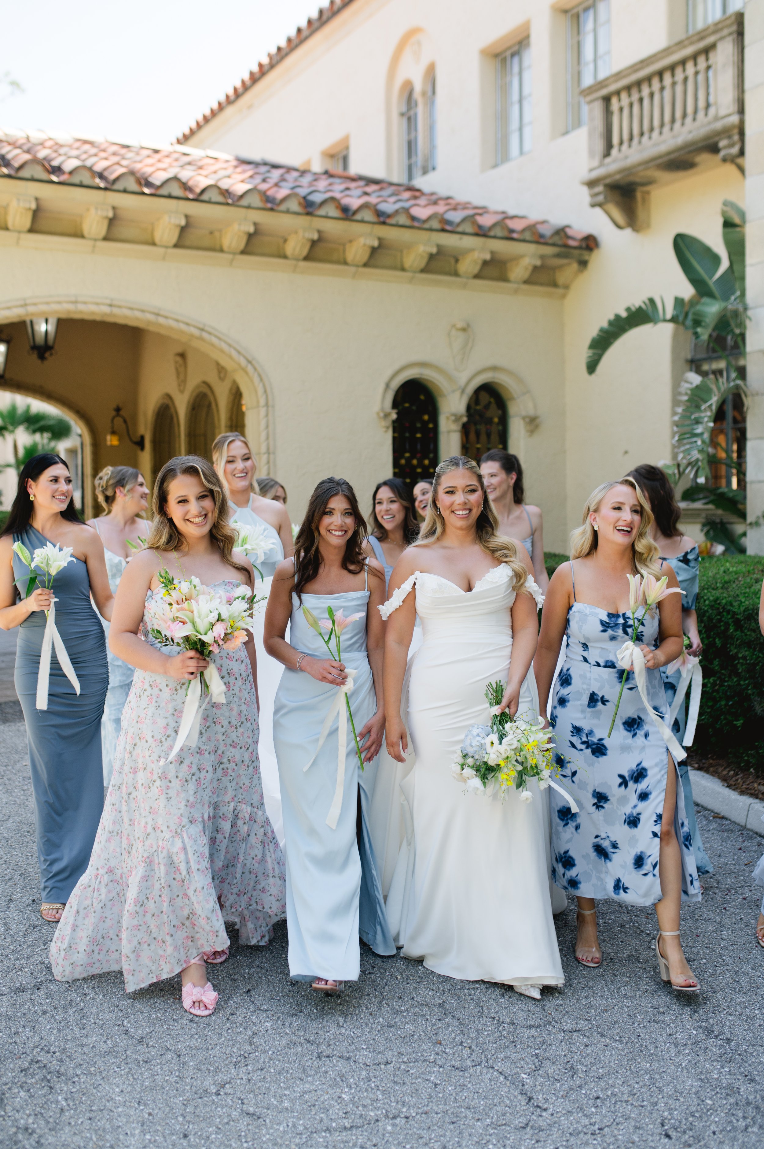 A group of women dressed in colorful dresses, holding bouquets of flowers, walking outside near a cream-colored building with a red-tiled roof.