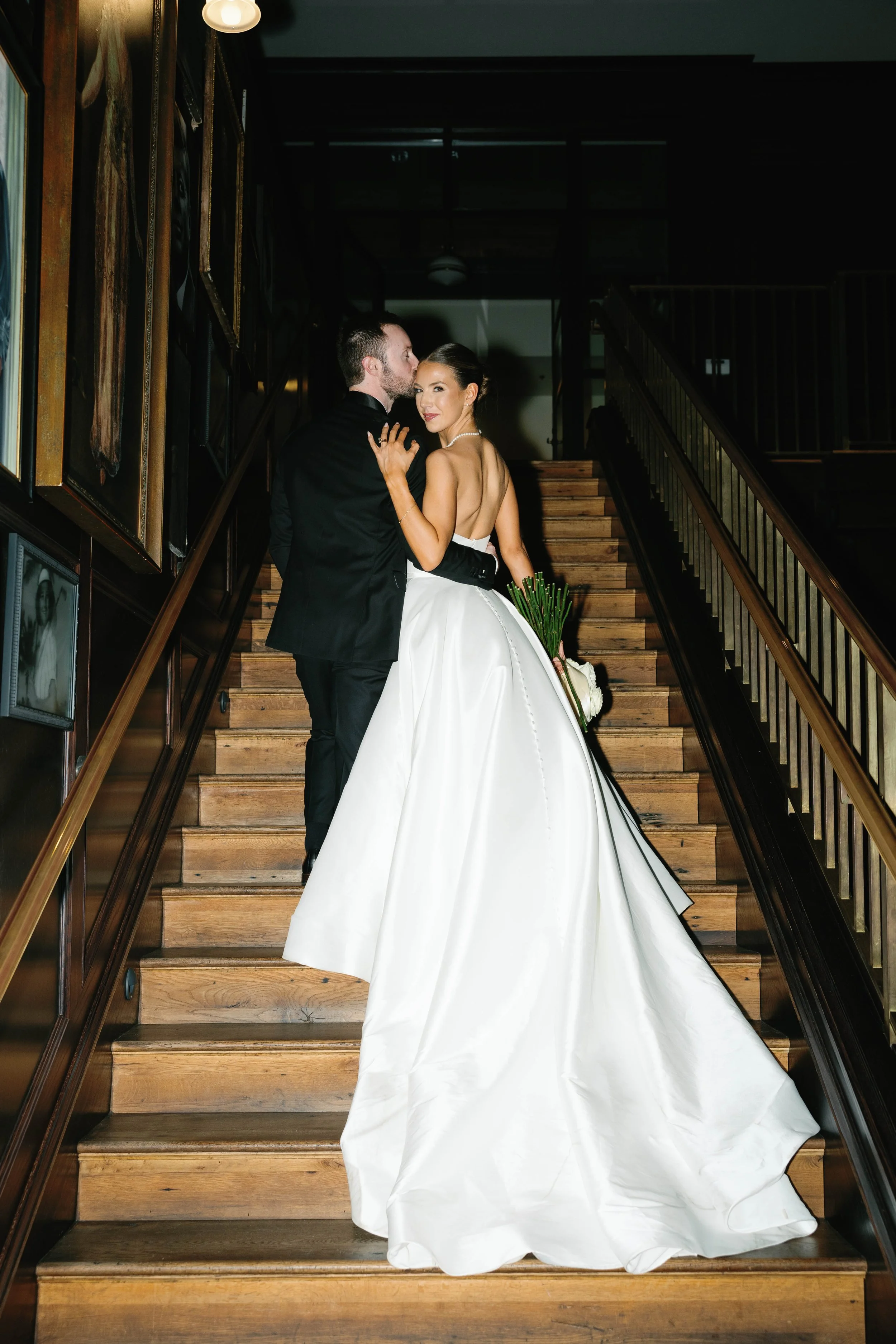 A bride and groom on a wooden staircase, the bride in a strapless white wedding gown holding a bouquet, the groom in a black tuxedo, in an elegant indoor setting with framed artwork on the walls.