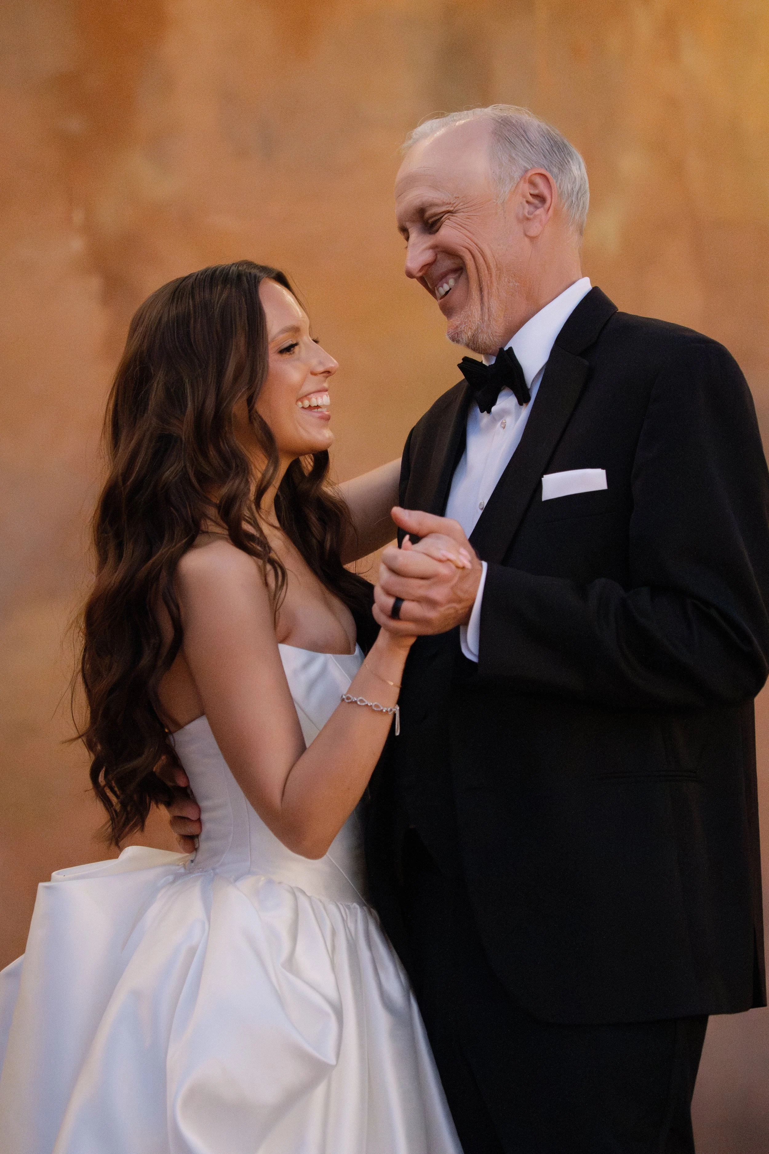 A young woman in a white wedding dress is dancing with an older man in a tuxedo, smiling at each other.