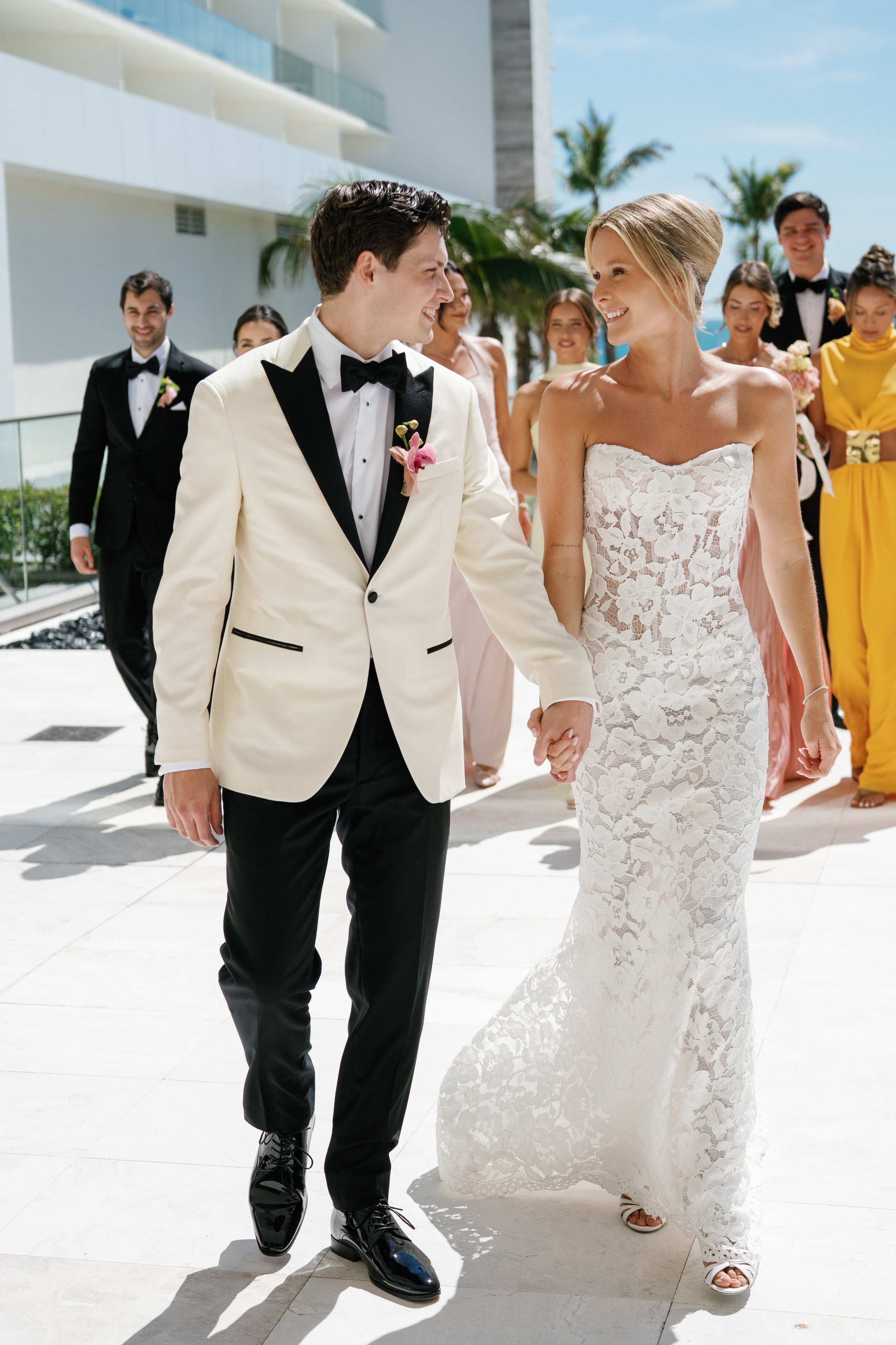 A couple dressed in wedding attire holding hands and smiling at each other, walking outdoors with a group of friends or family in the background near a modern building and palm trees on a sunny day.