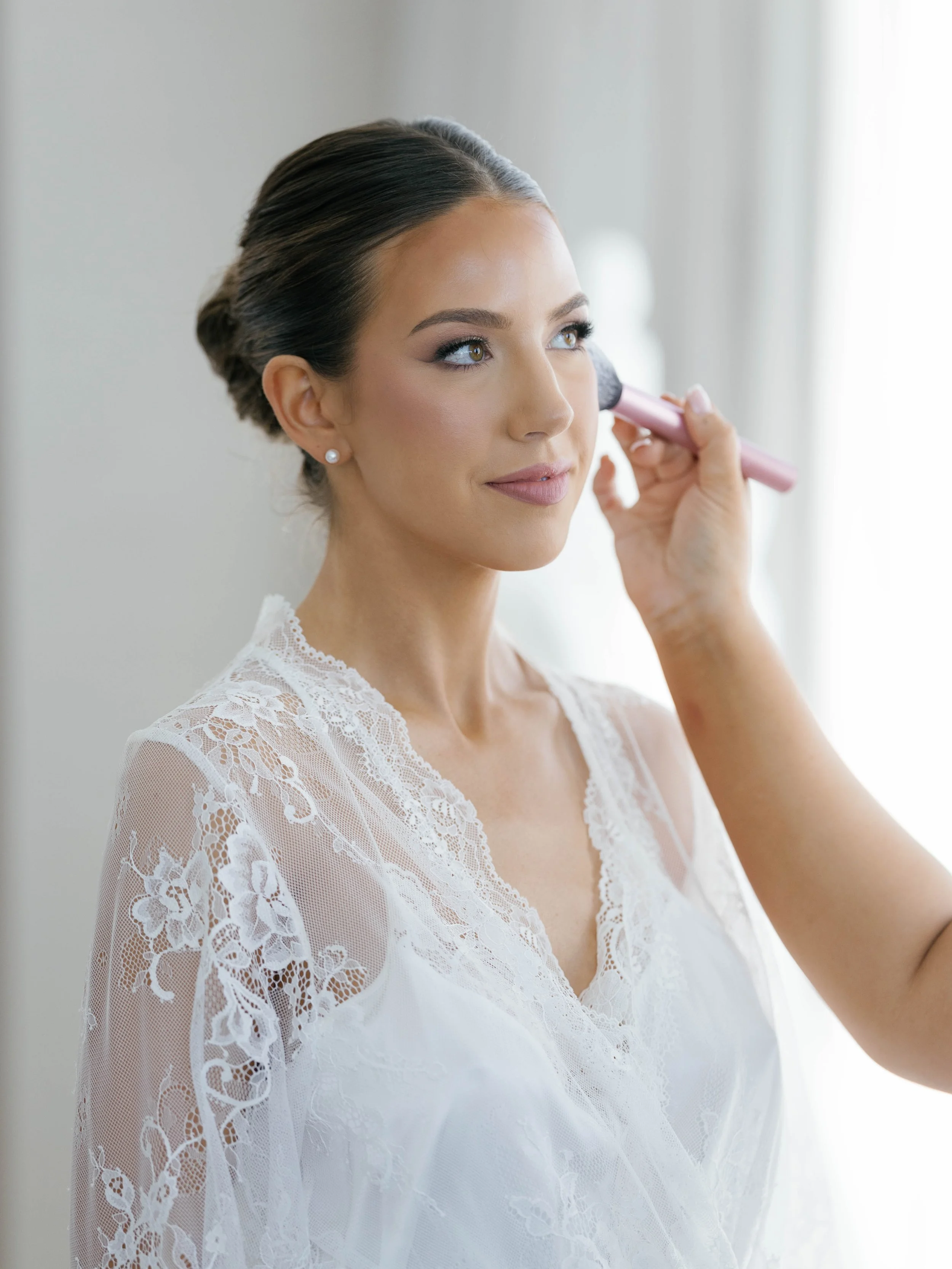 Woman with dark hair up, wearing pearl earrings and white lace dress, applying makeup with a brush.