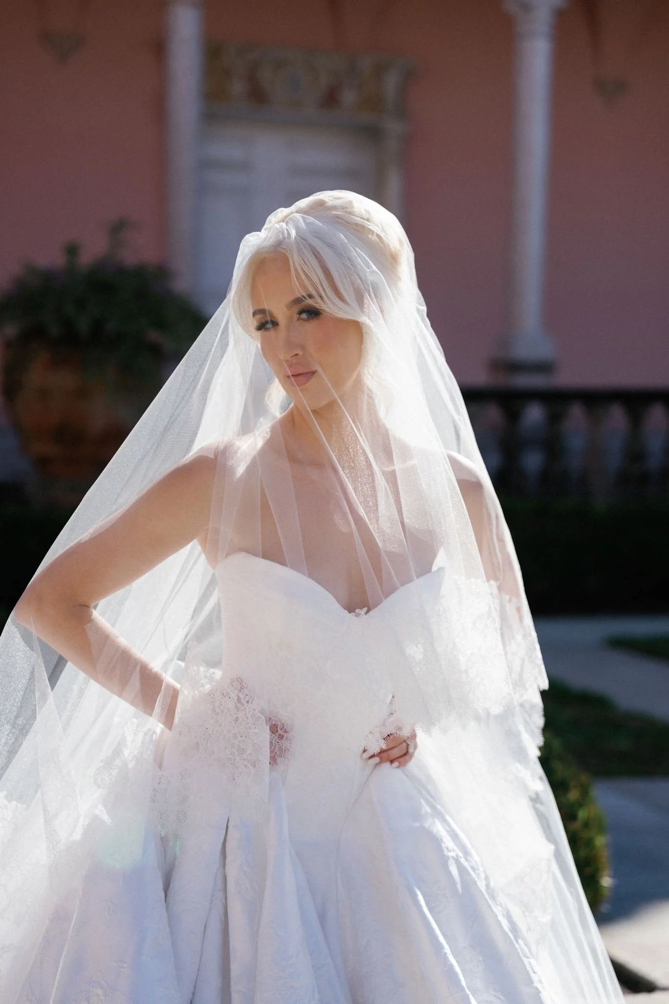 A bride in a white wedding gown and veil standing outdoors with a pink wall and a building in the background.