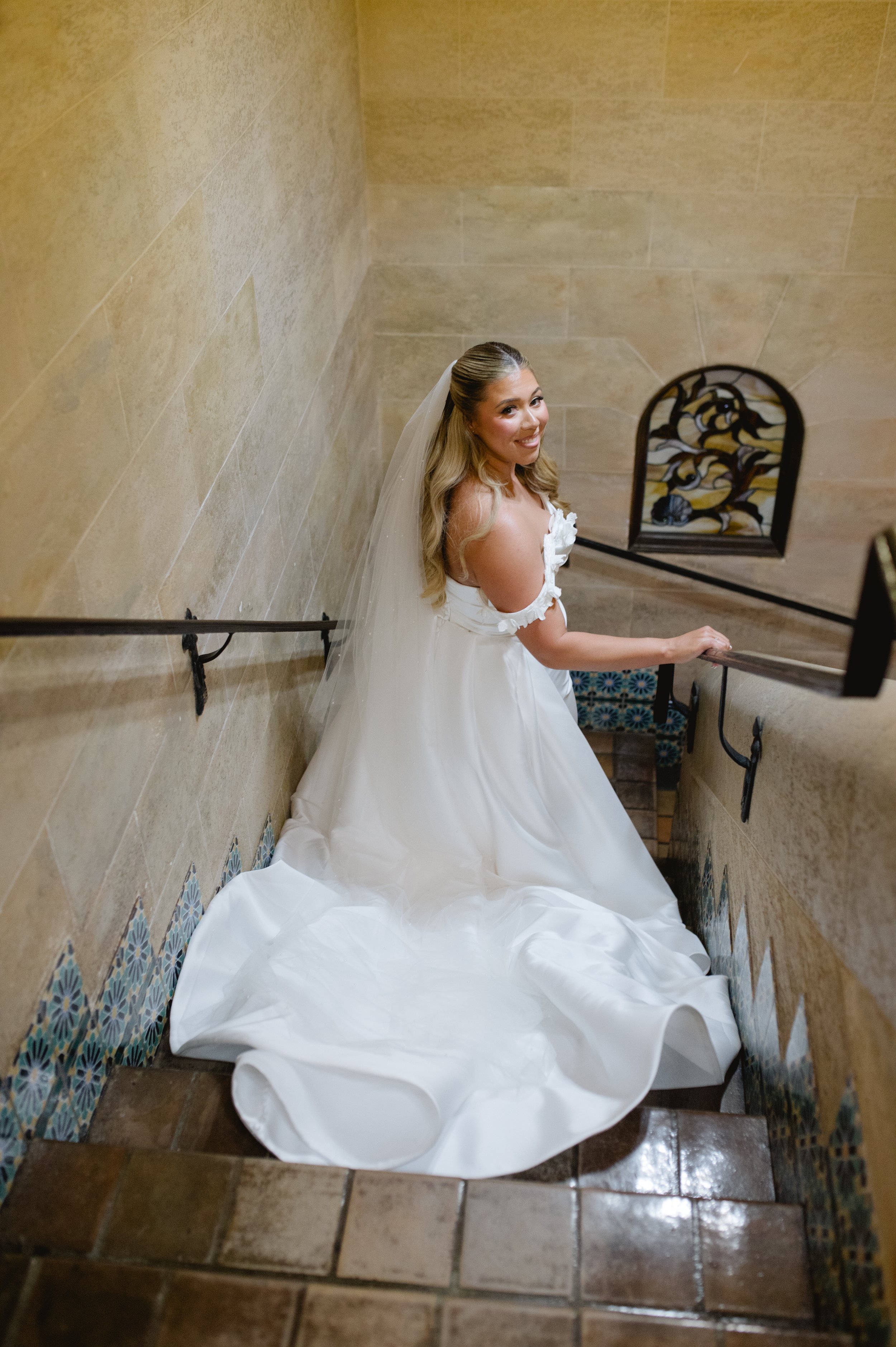 A smiling bride in a white wedding dress and veil sitting on a staircase with beige tile walls, holding onto a black iron railing.