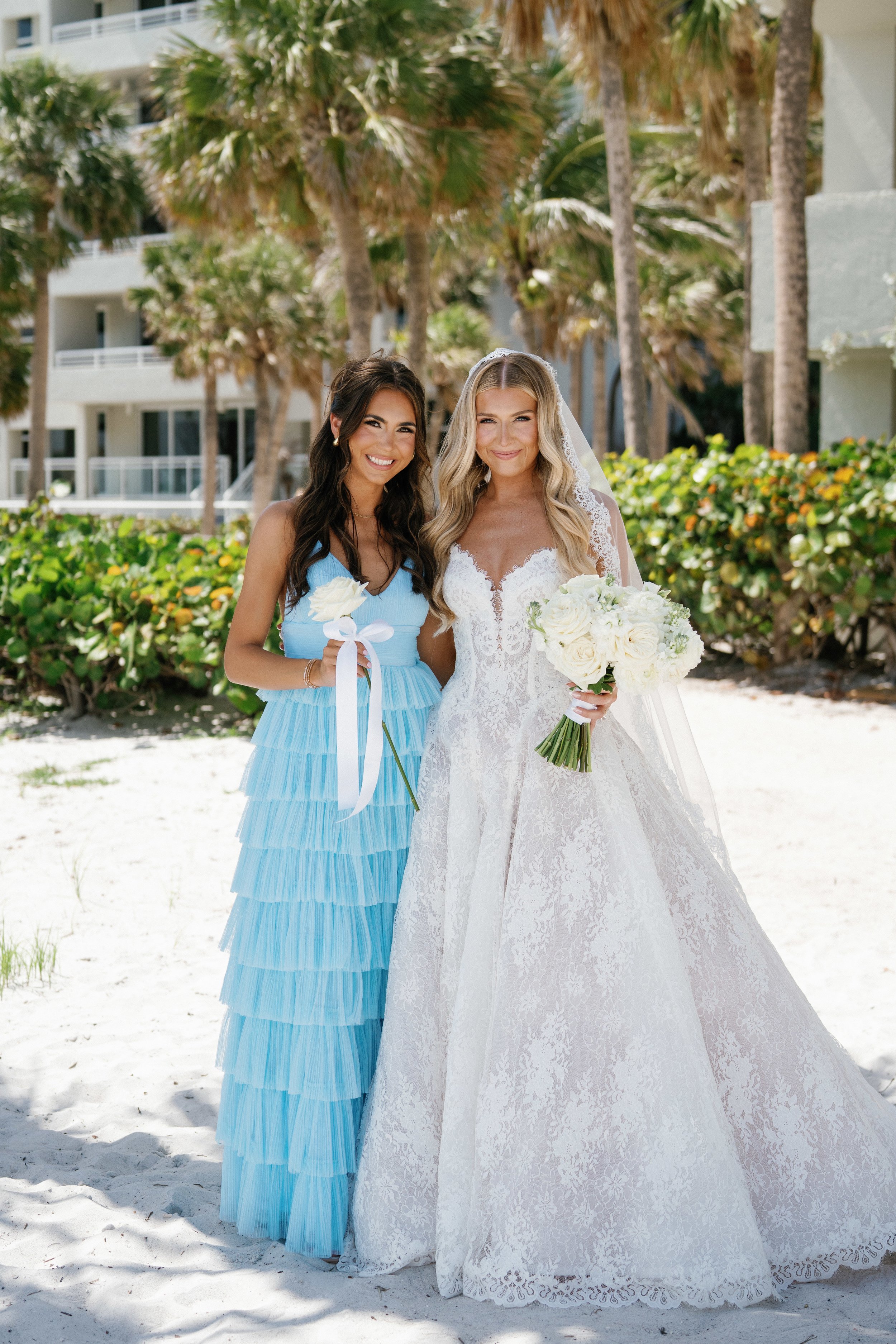 Two women standing on a beach, one in a white wedding dress with a bouquet of white roses, and the other in a tiered blue dress holding a white rose.