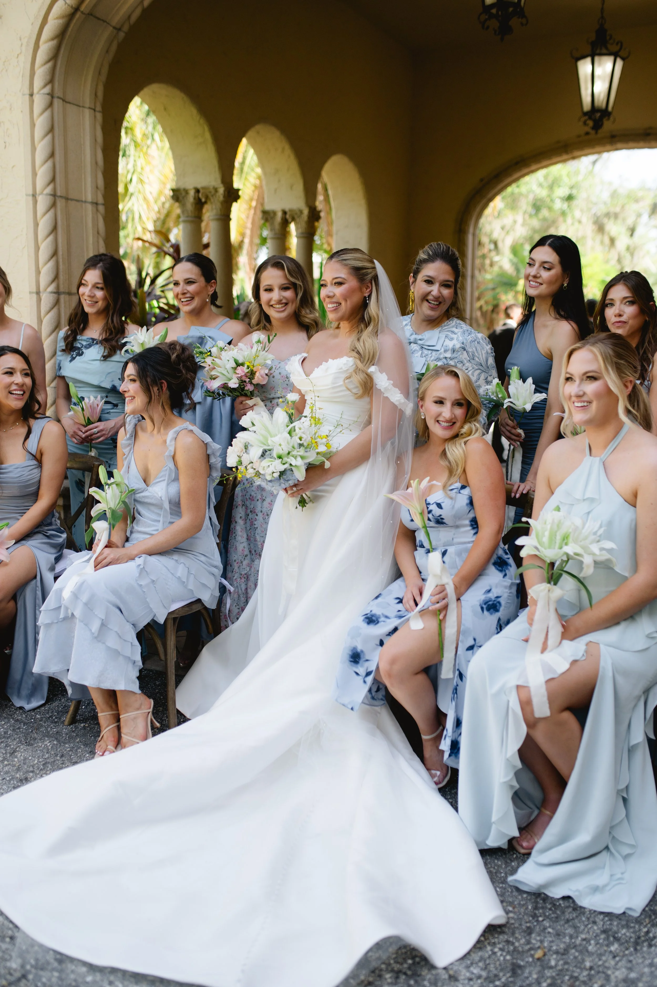 A bride in a white wedding gown holding a bouquet of white lilies and pink flowers stands among bridesmaids wearing blue and white dresses, in an outdoor archway with hanging lanterns and botanical scenery.